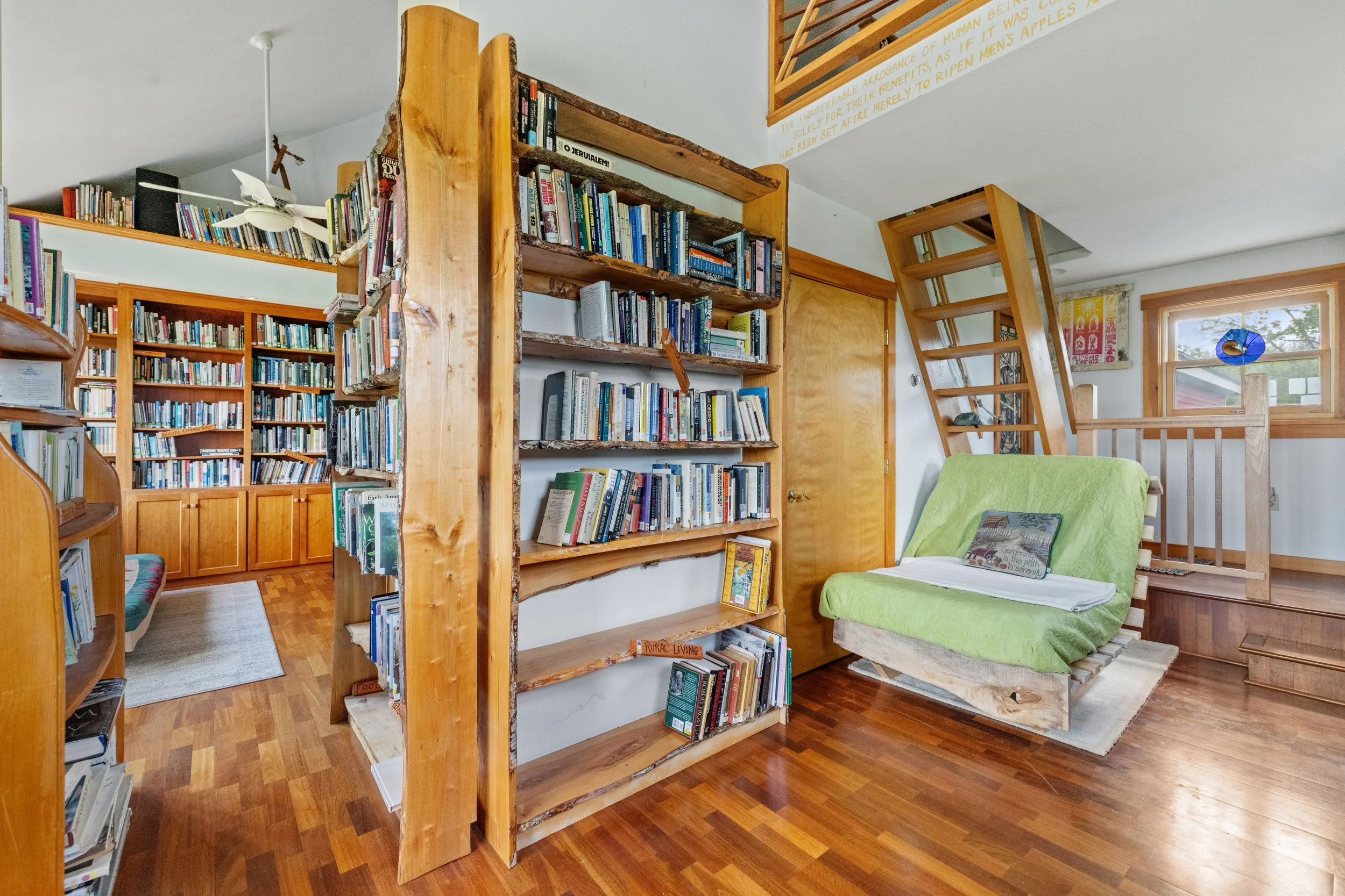 A cozy room with wooden floors and numerous bookshelves filled with books. There is a wooden staircase leading up to a loft. A green cushioned lounge chair or futon with a book on its armrest is positioned near a window. The room has a warm, inviting atmosphere with natural light coming through the windows.