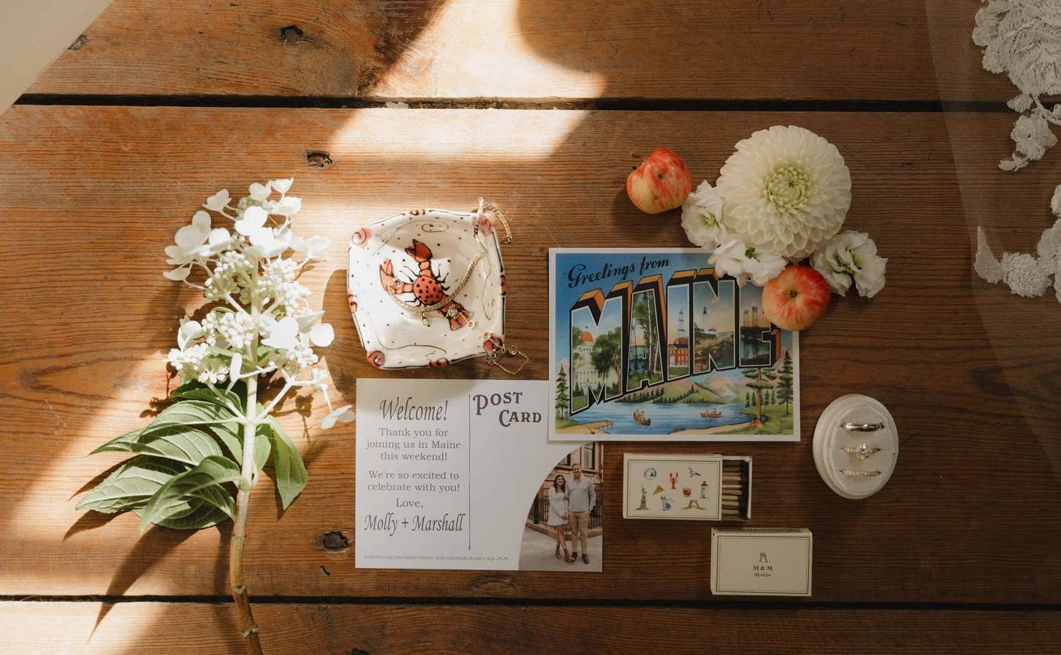 Flat lay of a rustic wooden table with a white flower branch, a postcard reading 'Greetings from Maine', three apples, a decorative bowl with a lobster design, white flowers, a welcome card for an event in Maine, a small matchbox, and a jewelry ring