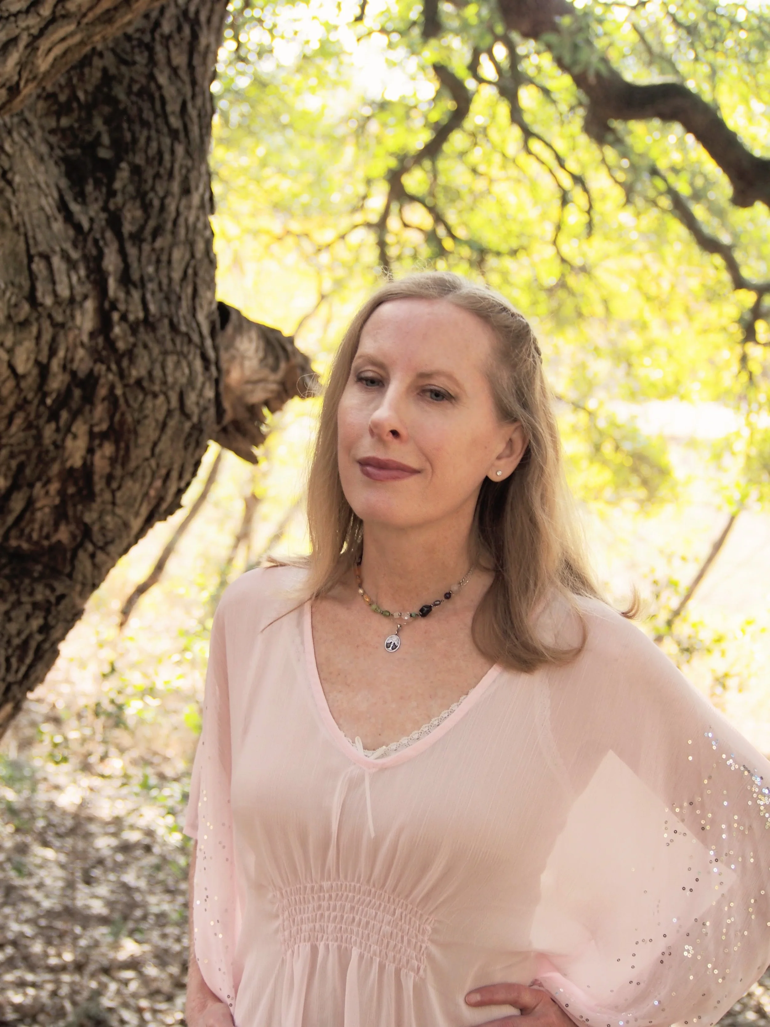 A woman with blonde hair standing outdoors near a large tree with green leaves in the background. She is wearing a light pink top with a gemstone beaded necklace and has a calm, meditative and confident expression.