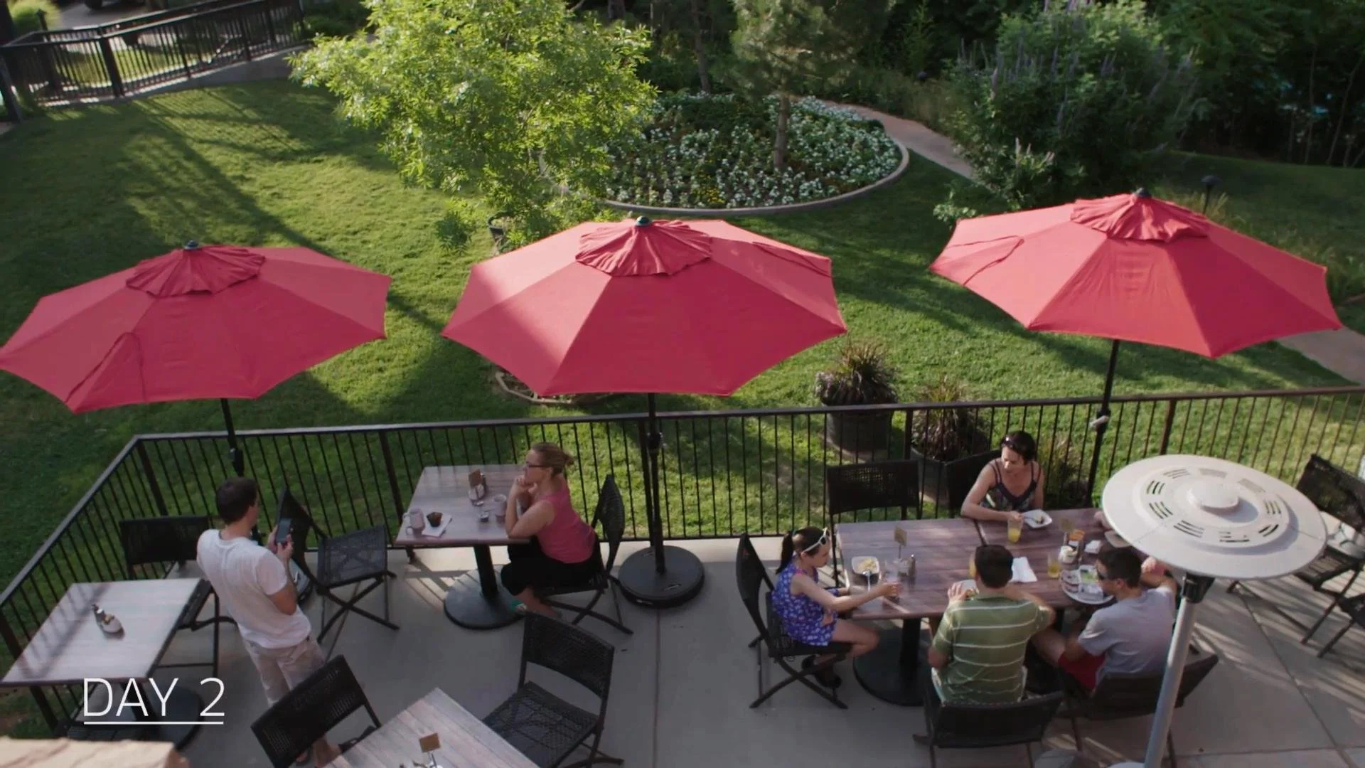 Outdoor patio with people dining under pink umbrellas. There is a grassy backyard with a circular flower bed and a patio railing.