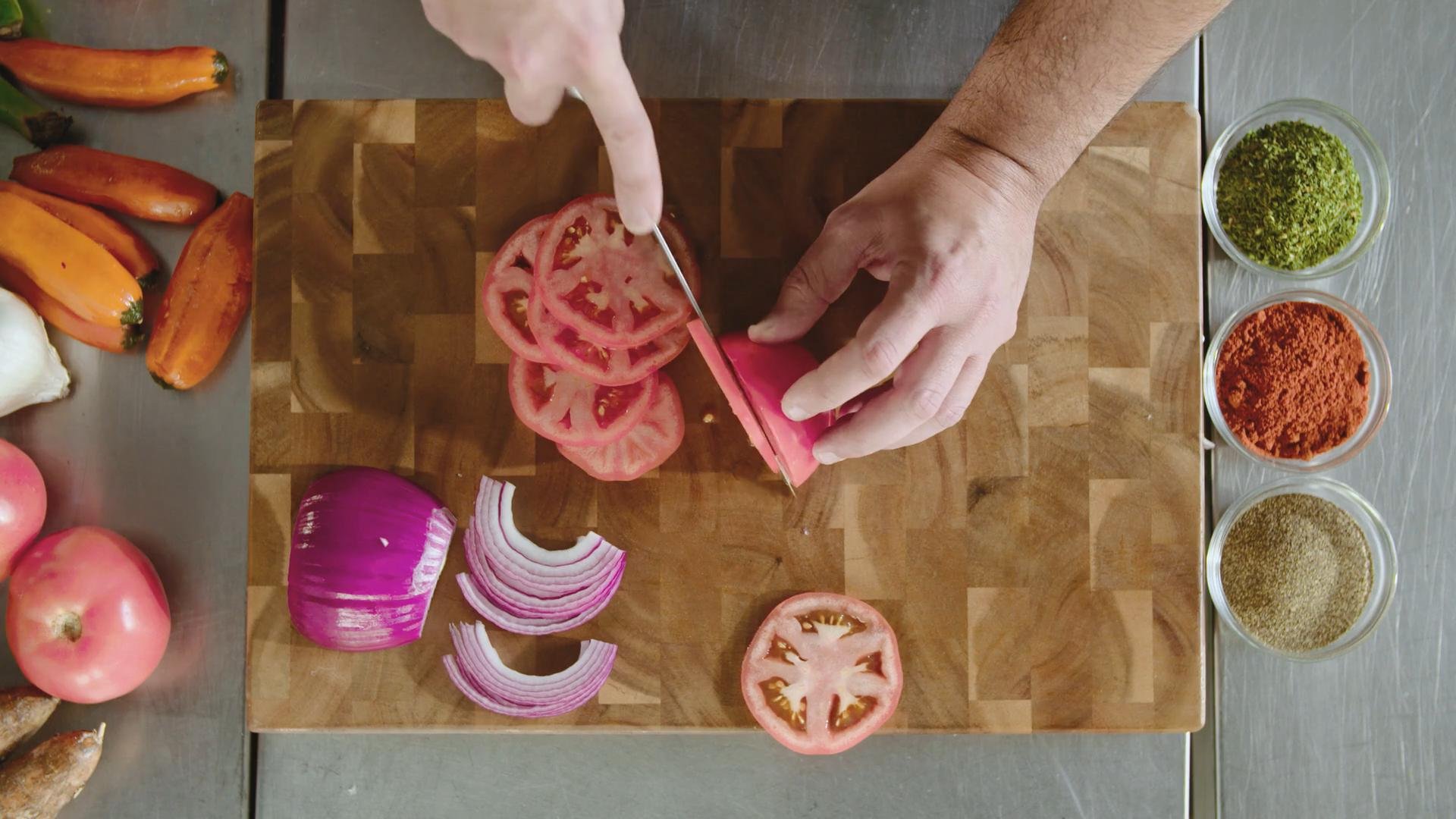 Person slicing tomatoes on a wooden cutting board with sliced onion nearby and bowls of chopped herbs and spices arranged on the side.