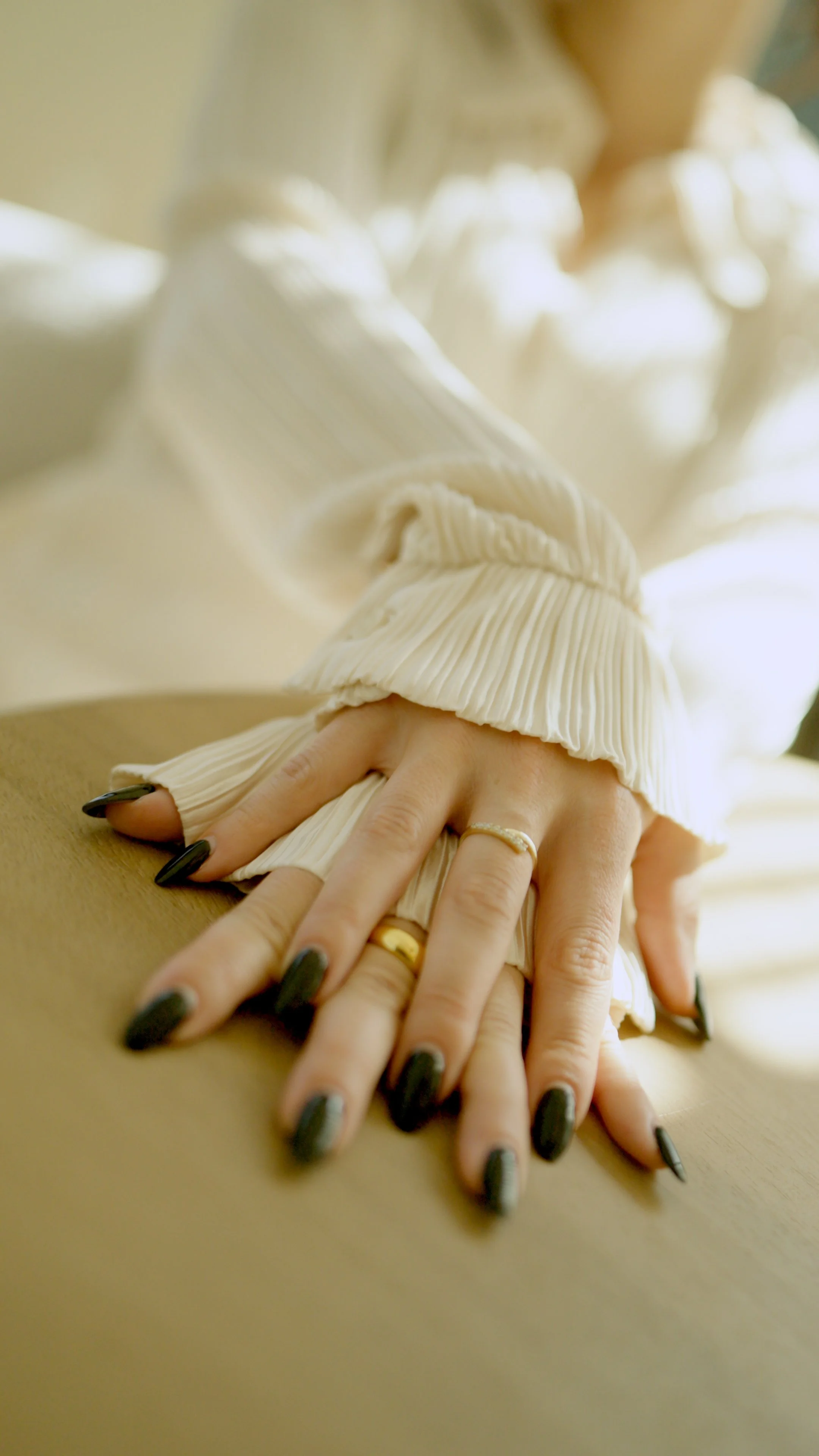 Close-up of a person's hand resting on a flat surface, with black painted nails, wearing multiple rings, with the sleeve of a beige or cream-colored pleated fabric visible.