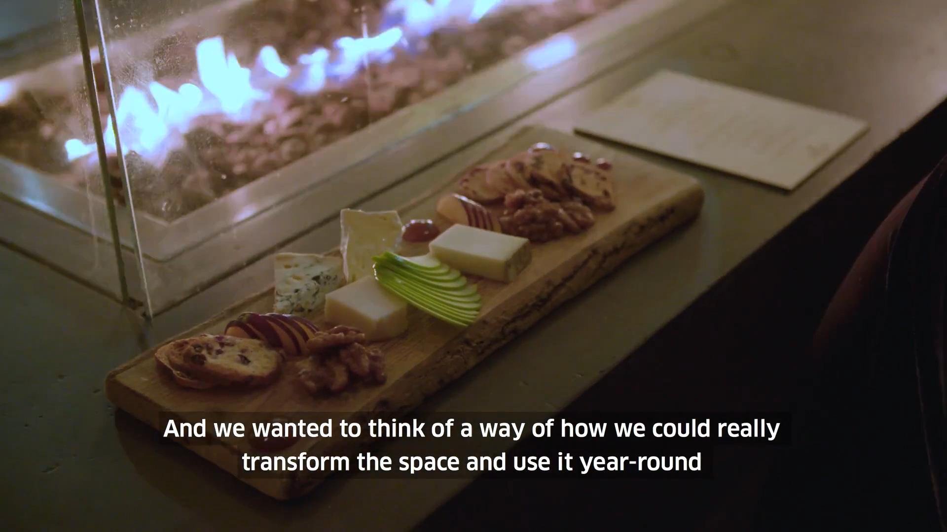 A cheese and charcuterie board with various cheeses, meats, sliced apples, and garnishes, placed near a glowing fire pit.