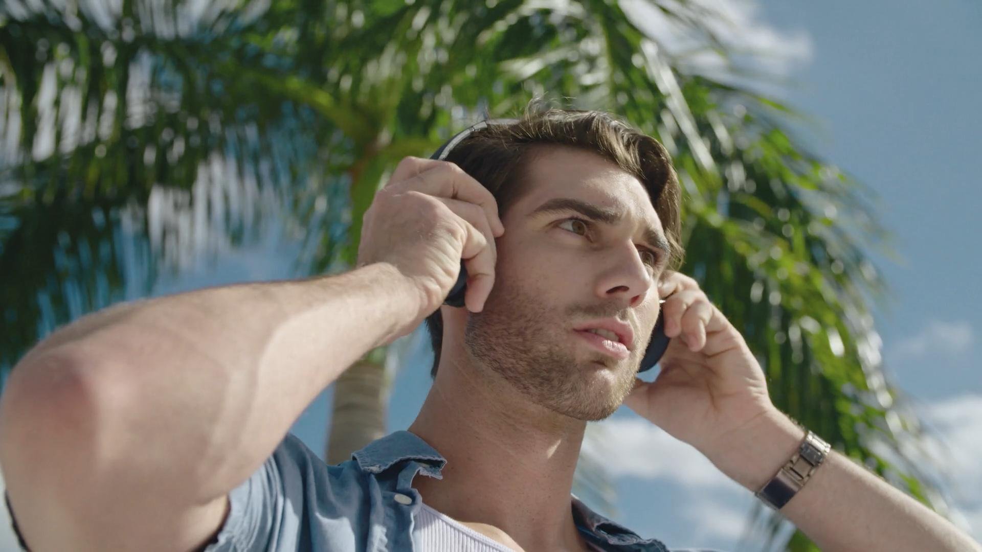 Young man with light skin and brown hair, outdoors, adjusting headphones against a background of green palm leaves and blue sky.
