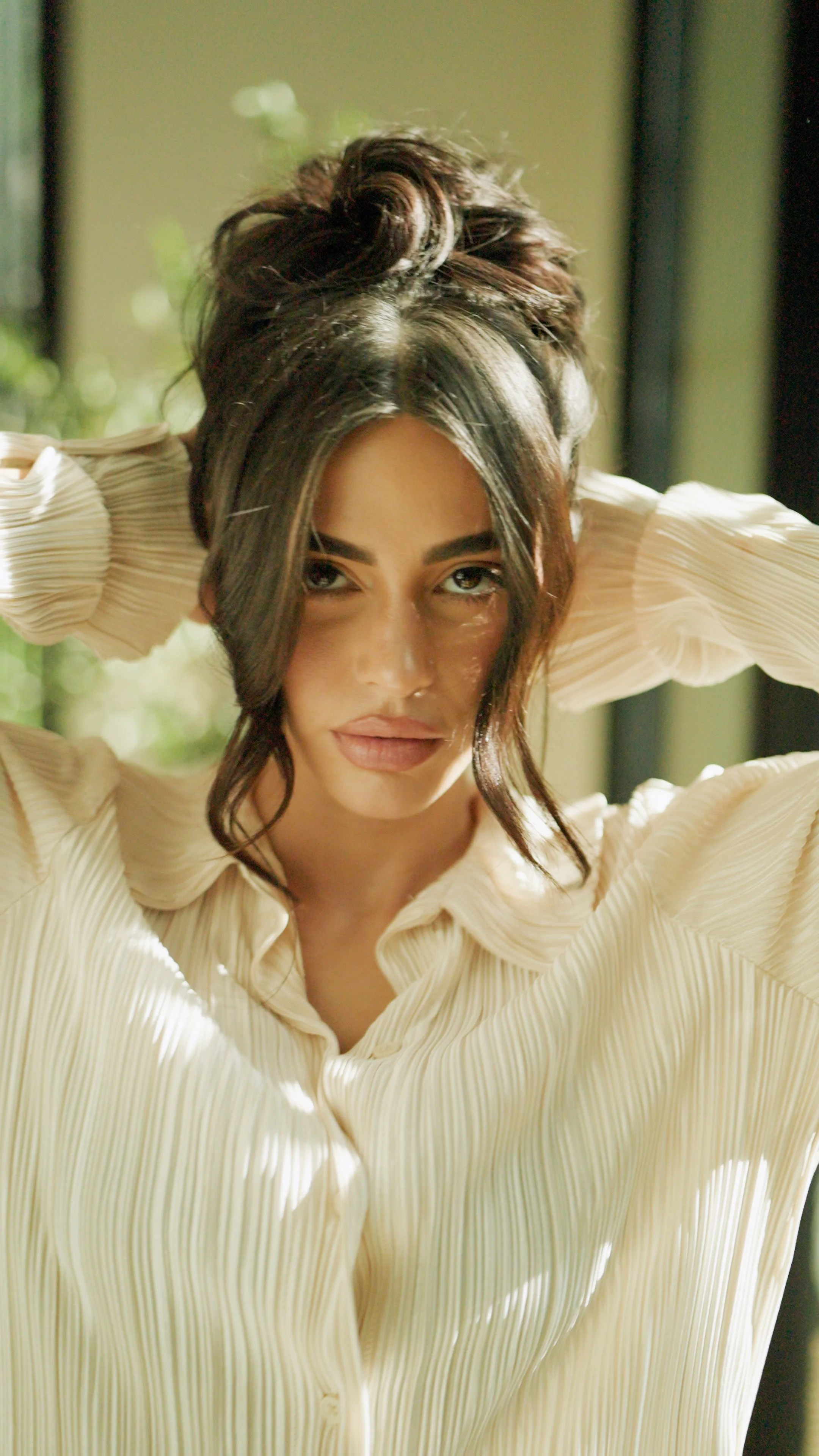 A woman with dark hair styled in a bun, wearing a cream-colored, pleated blouse, looking directly at the camera with her hands behind her head.