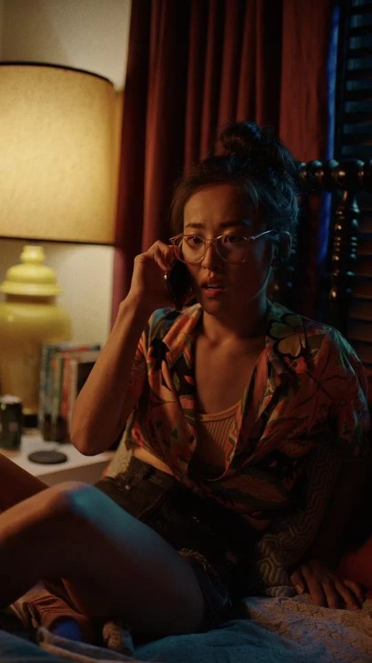 A woman with glasses and a top knot hairstyle is sitting on a bed, speaking on her cell phone, in a dimly lit room with a large lamp, books, and a dark wooden headboard in the background.