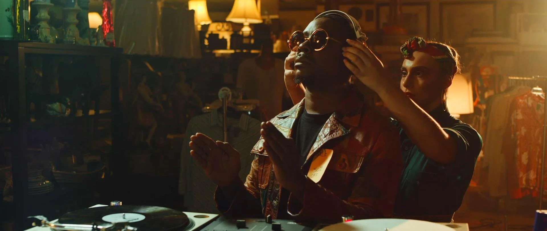 A DJ is adjusting headphones on a man in a dimly lit room. The man is wearing sunglasses and a patterned jacket, standing in front of a turntable with other people and vintage decor in the background.