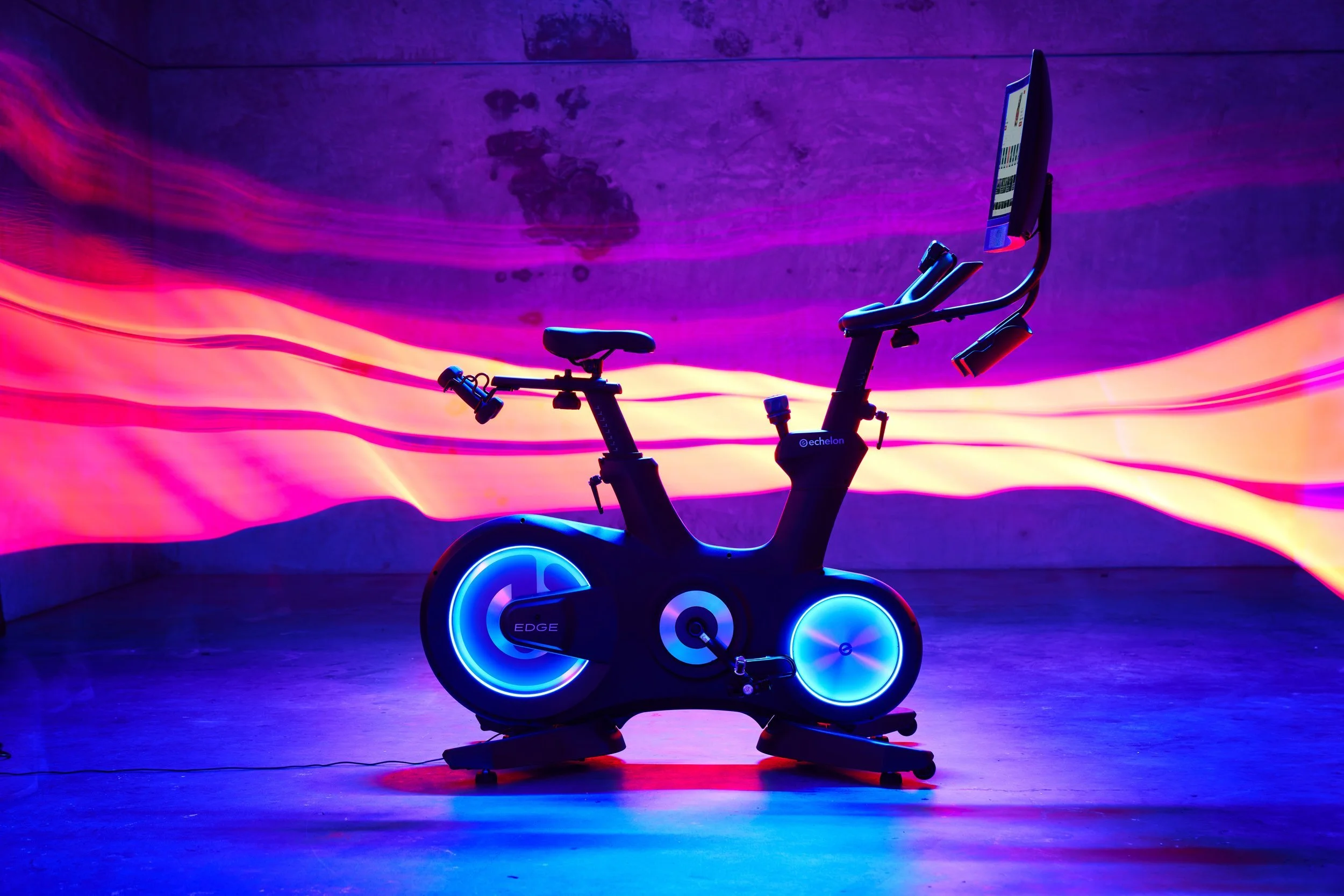 A stationary exercise bike illuminated with vibrant blue and pink lights against a dark wall with colorful light trails.