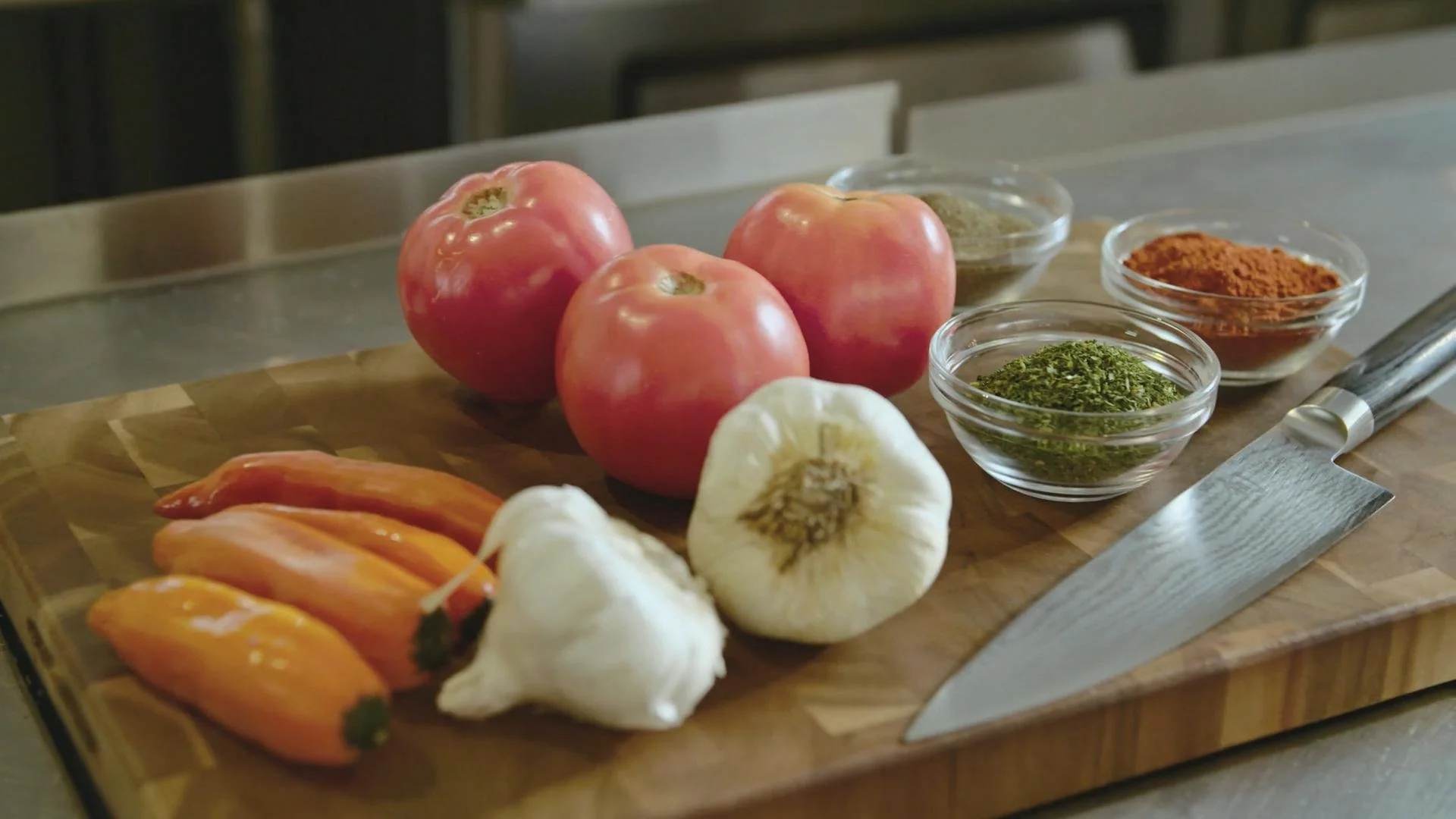 Fresh tomatoes, garlic, peppers, and bowls of spices on a wooden cutting board with a chef's knife.