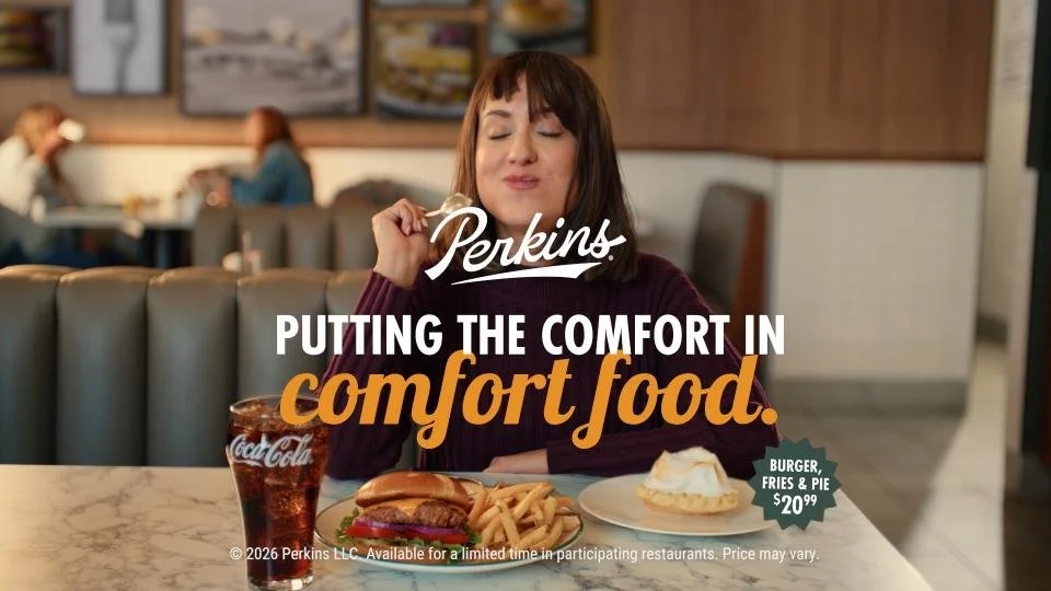 A woman sitting in a restaurant enjoying comfort food, with a cheeseburger, fries, pie, and a Coca-Cola on the table; the scene advertises Perkins restaurant and its comfort food.