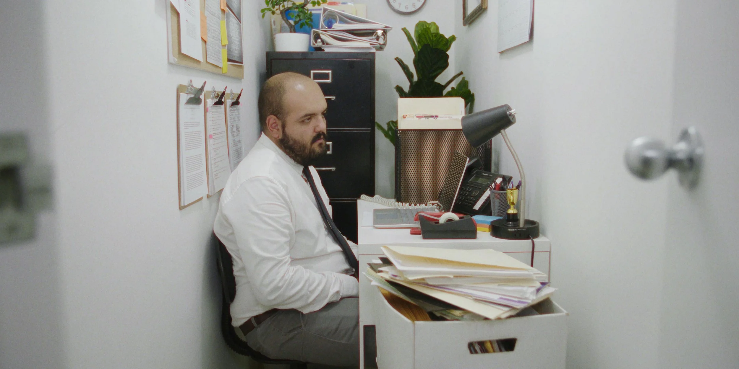 A man sits at a cluttered desk in a small office, wearing a white shirt and tie, surrounded by documents, a lamp, a phone, and a filing cabinet, with a bulletin board and a plant on the wall behind him.