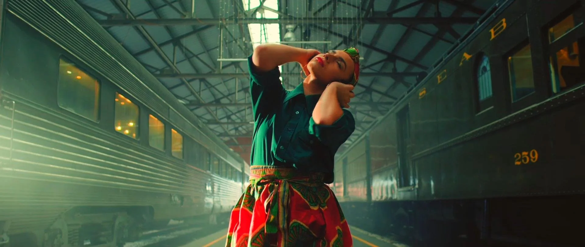 A woman with colorful hair and red lipstick poses inside a train station, wearing a green shirt and a multicolored skirt. The station has high arches, and there are trains on either side of her.