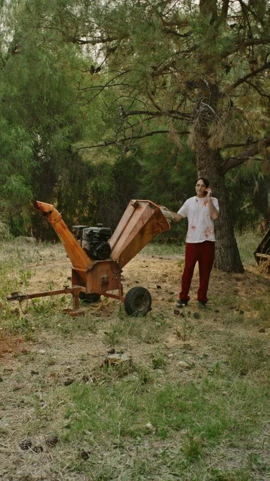 A woman dressed like a zombie with fake blood stains on her white shirt and red pants, standing outdoors in a wooded area, next to a rusty wood chipper machine.