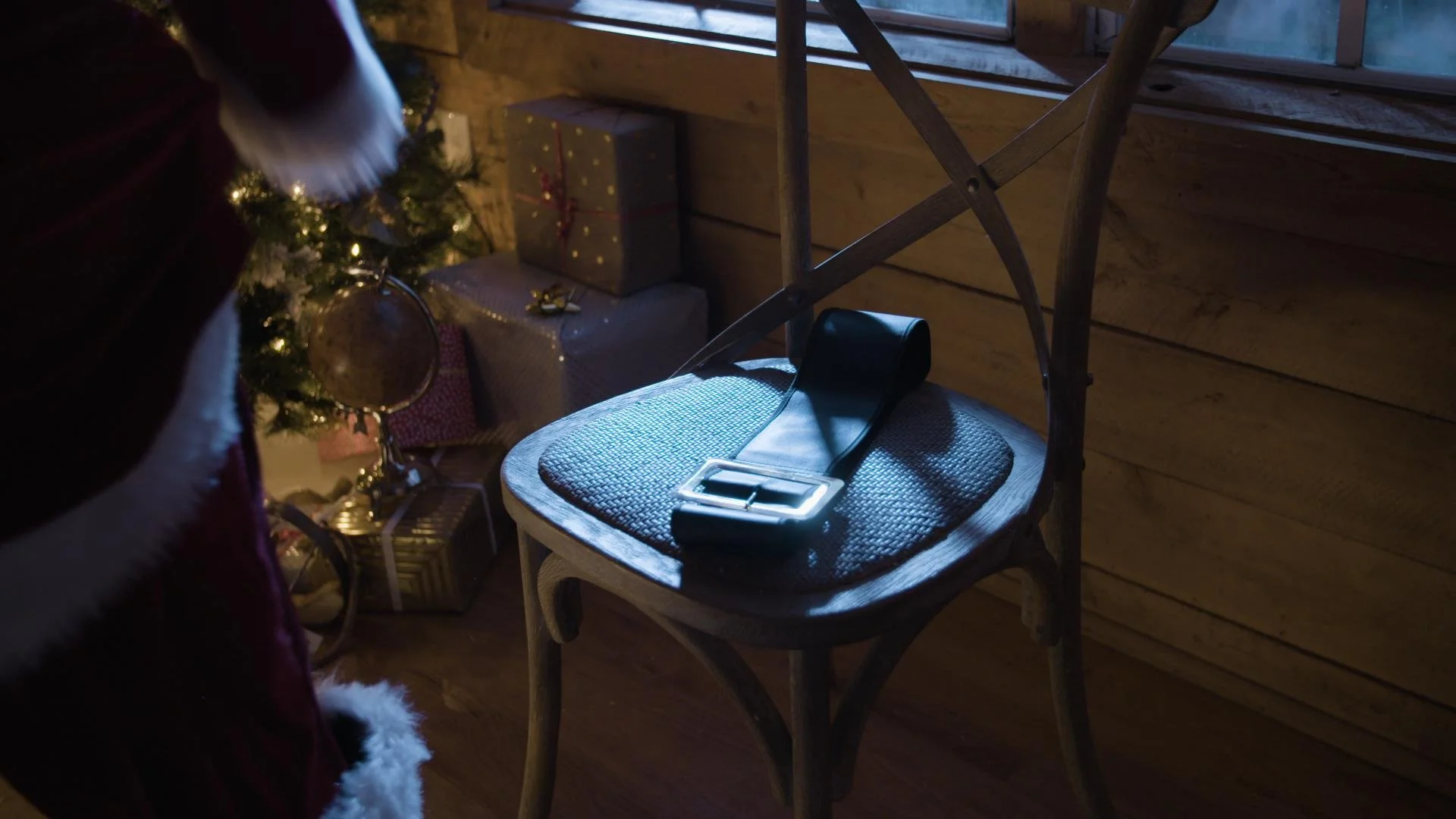 Wooden chair with a black weightlifting belt and a smartphone on it, in a cozy room with wooden walls, decorated for Christmas with a small decorated Christmas tree and wrapped presents.