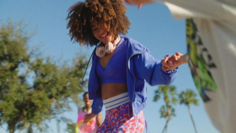 A woman with curly hair, wearing a blue sports bra, a blue jacket, and colorful shorts, smiling and holding hands with someone outside on a sunny day with trees in the background.