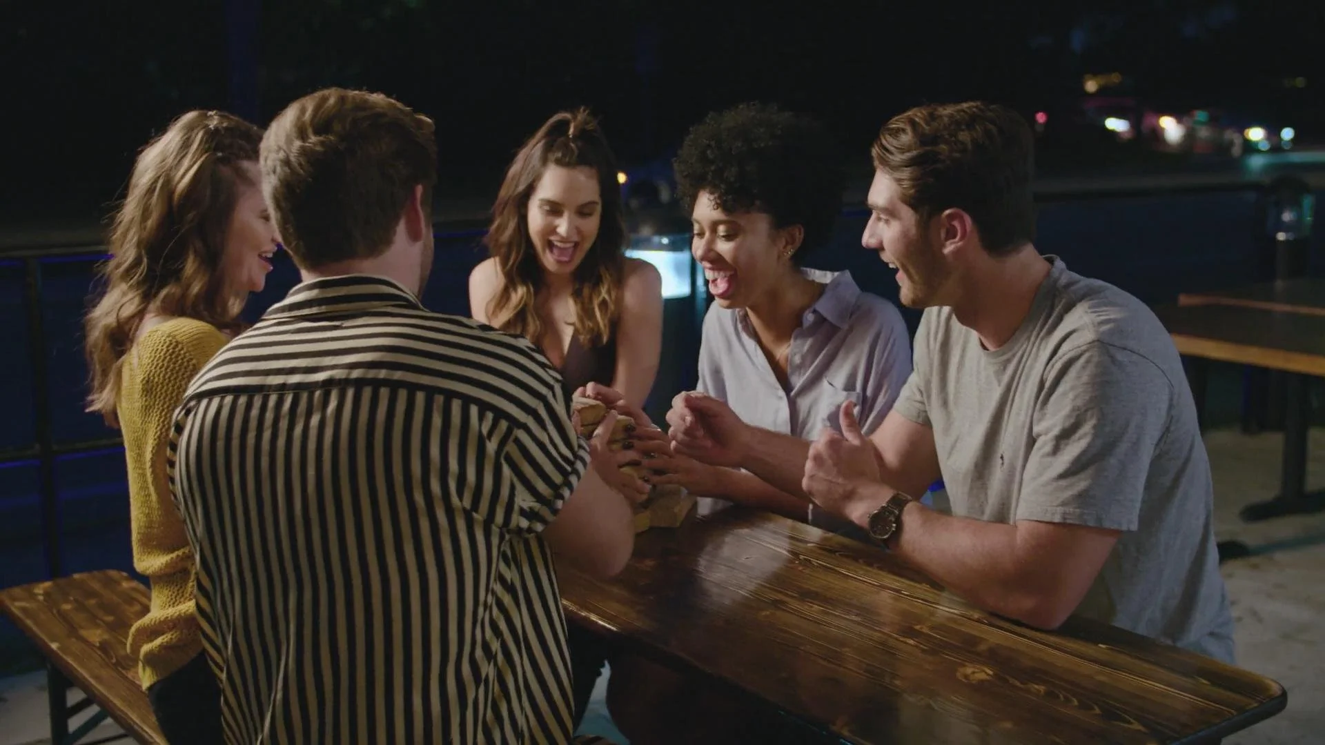 Six young adults sitting around a wooden table outdoors at night, smiling and looking at a phone they are holding together.