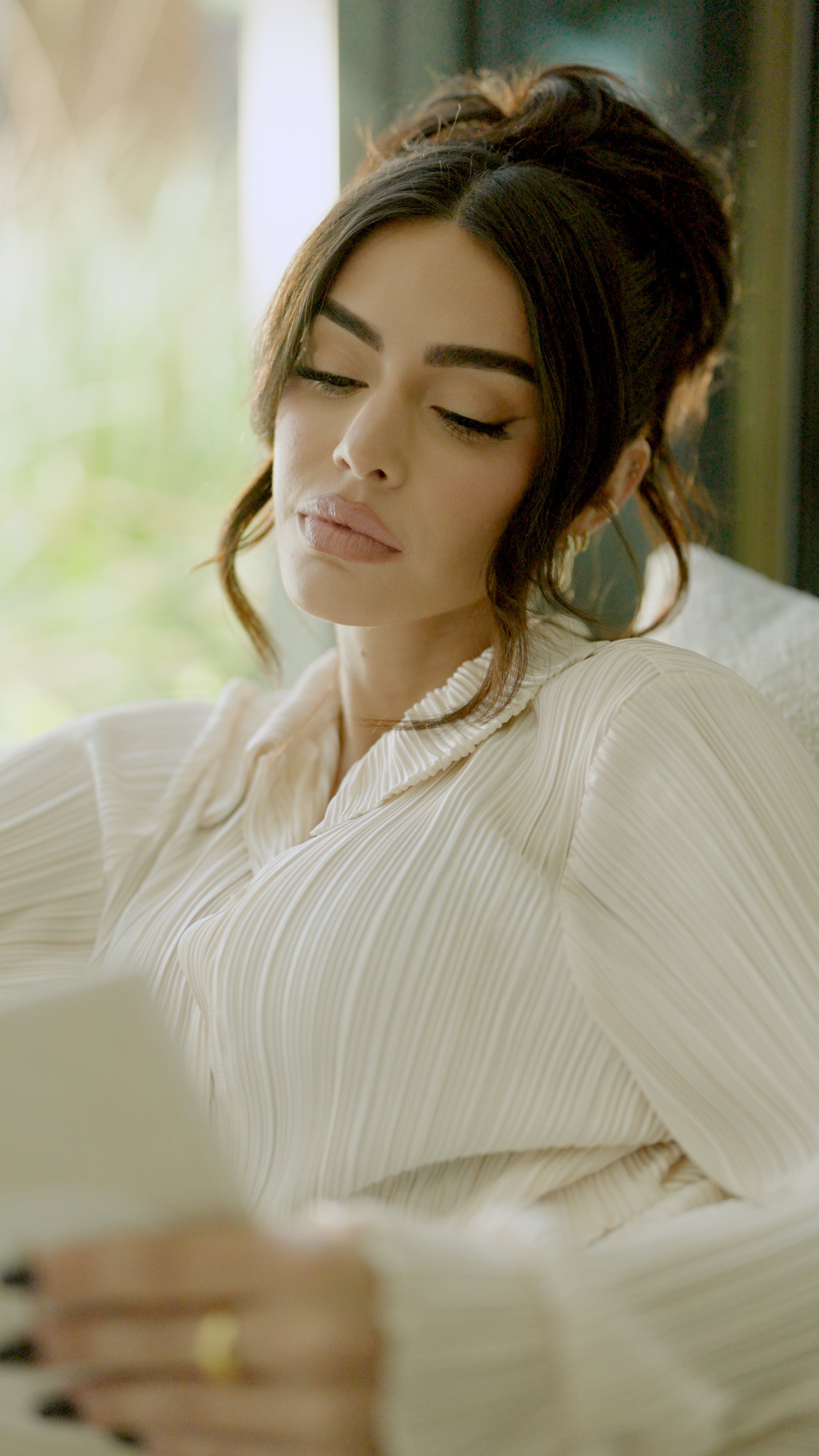 A woman with dark hair styled in a messy bun, wearing a cream-colored, pleated blouse, looking down, sitting near a window with blurred green outdoor scenery in the background.