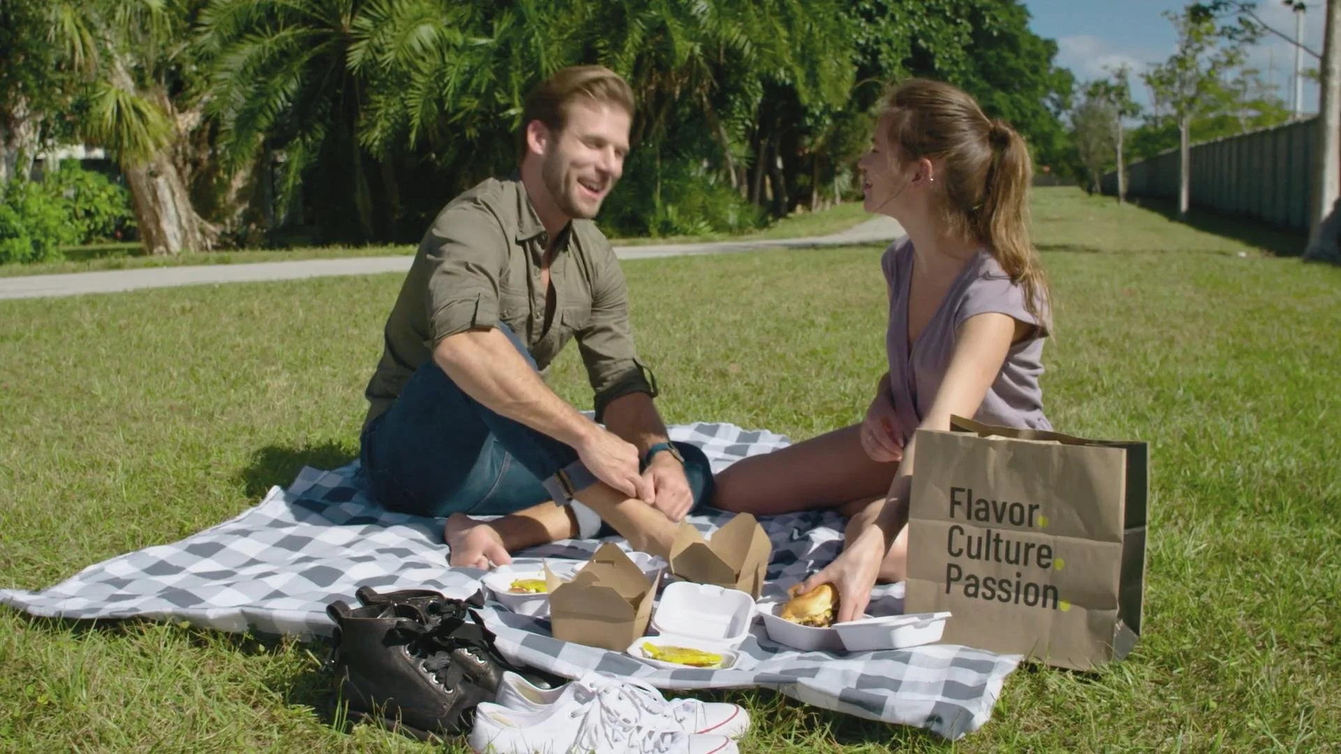 A couple sitting on a picnic blanket in a park, sharing food and smiling at each other, with shopping bags nearby.