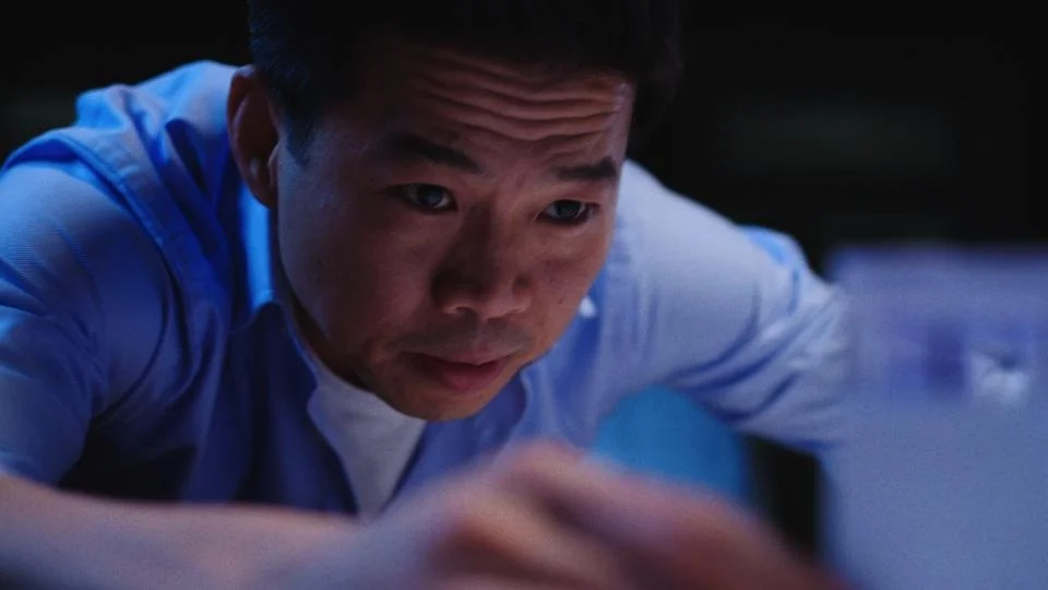 A young man with short black hair in a blue shirt is looking down at a device, with a focused expression, in a dimly lit environment.