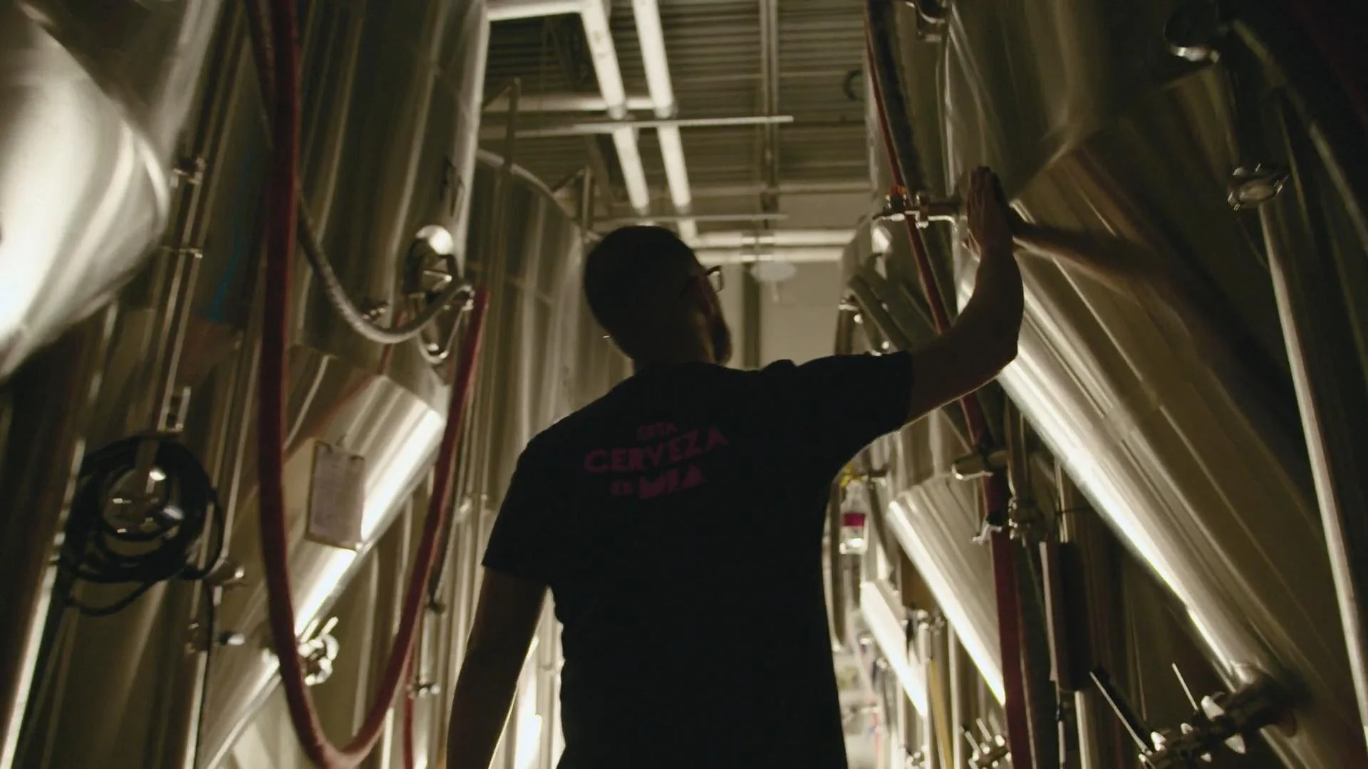 A person wearing glasses and a dark T-shirt with pink text on the back, reaching up to inspect or adjust equipment in an industrial setting with large stainless steel tanks and piping.