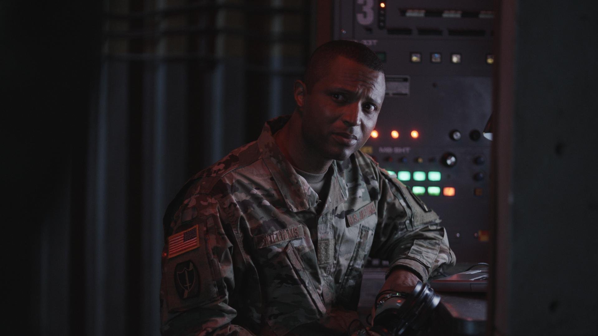 A man in military uniform sitting at a control panel in a dimly lit room, looking at the camera.
