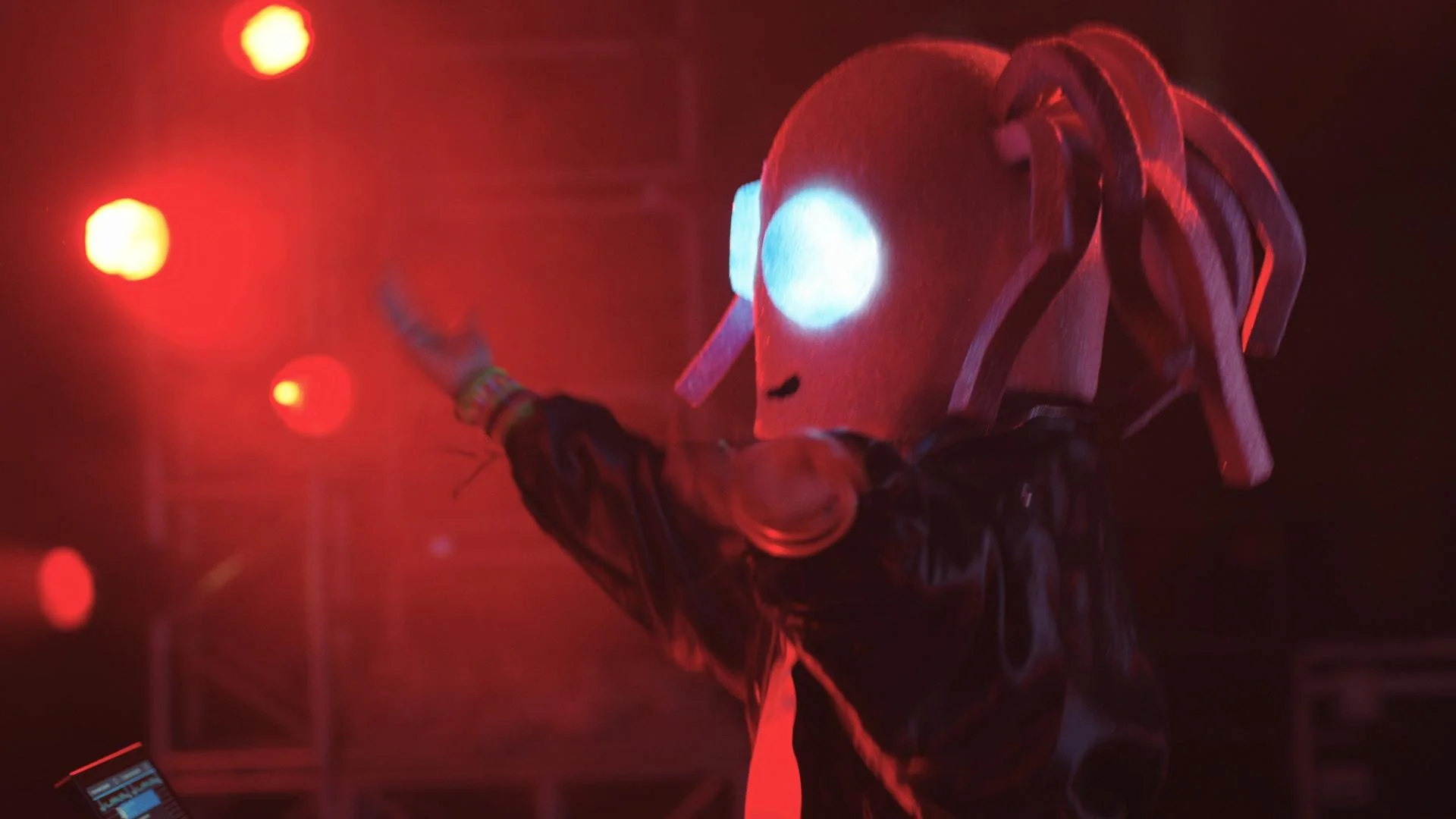 A performer wears a white astronaut helmet with glowing blue light in a dark setting illuminated by red lights, with a hand raised and holding an object.
