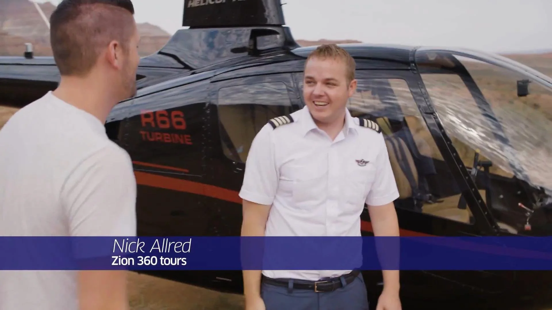 Two men standing outside near a helicopter. One man facing away wearing a white shirt, the other man smiling wearing a pilot uniform. The helicopter is black with red and orange accents and has the text 'R66 TURBINE' on the side. A landscape of open 