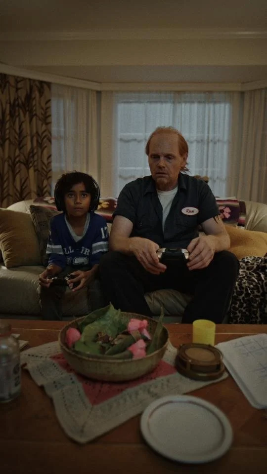 A man and a young girl sitting on a couch, playing video games in a living room with a window in the background.