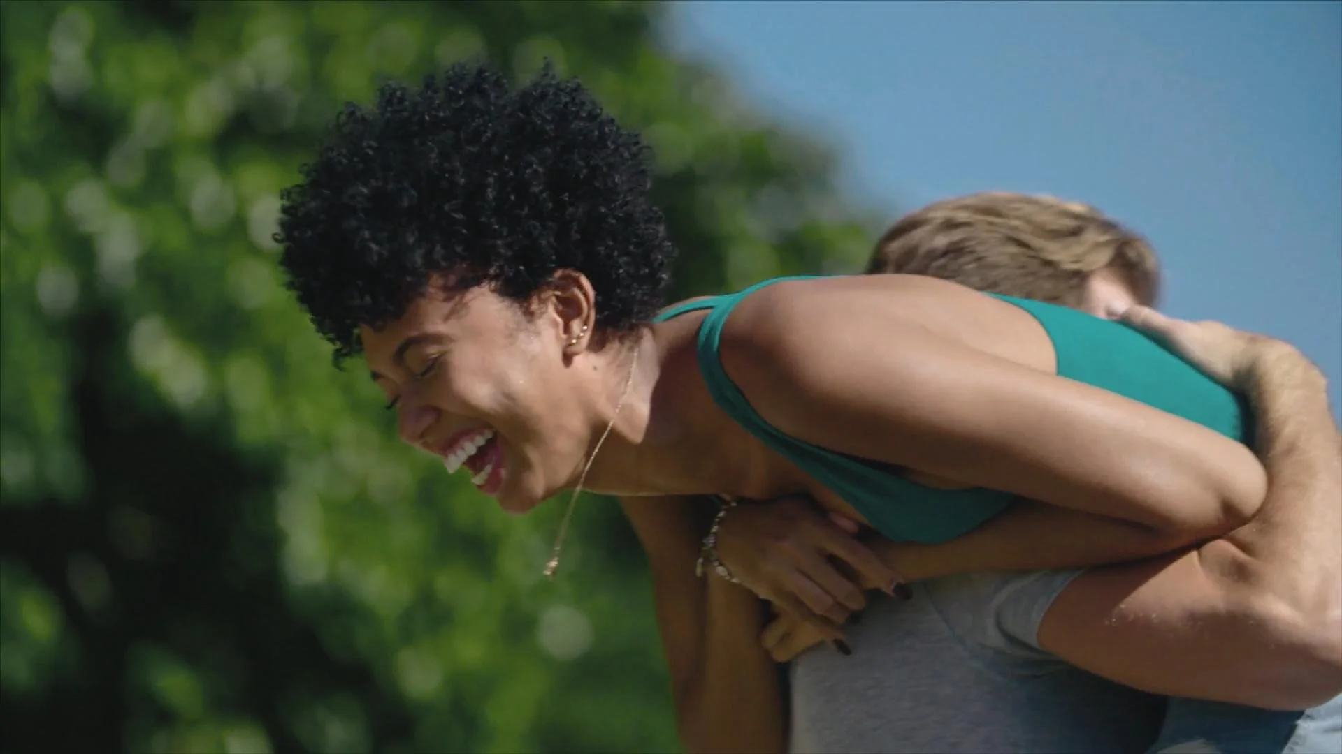 Two women engaged in an outdoor activity, with one woman smiling and holding onto the other from behind, both wearing athletic clothing, in a green outdoor setting.