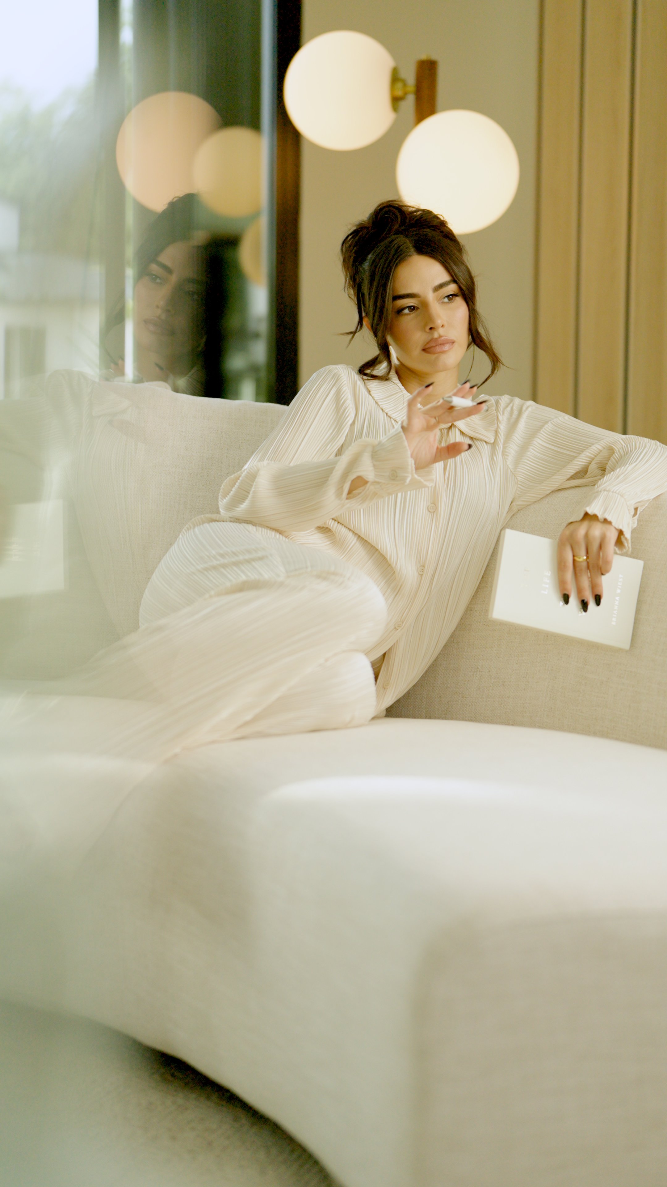 A woman with dark hair styled in a loose updo is reclining on a beige sofa in a modern living room, holding a book and a cigarette, with a thoughtful expression. There are three spherical modern ceiling lights above her and a glass window reflecting 