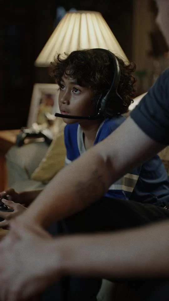 A young boy with curly hair wearing a game headset and holding a game controller, sitting on a couch in a dimly lit living room with a lamp and picture frame in the background.