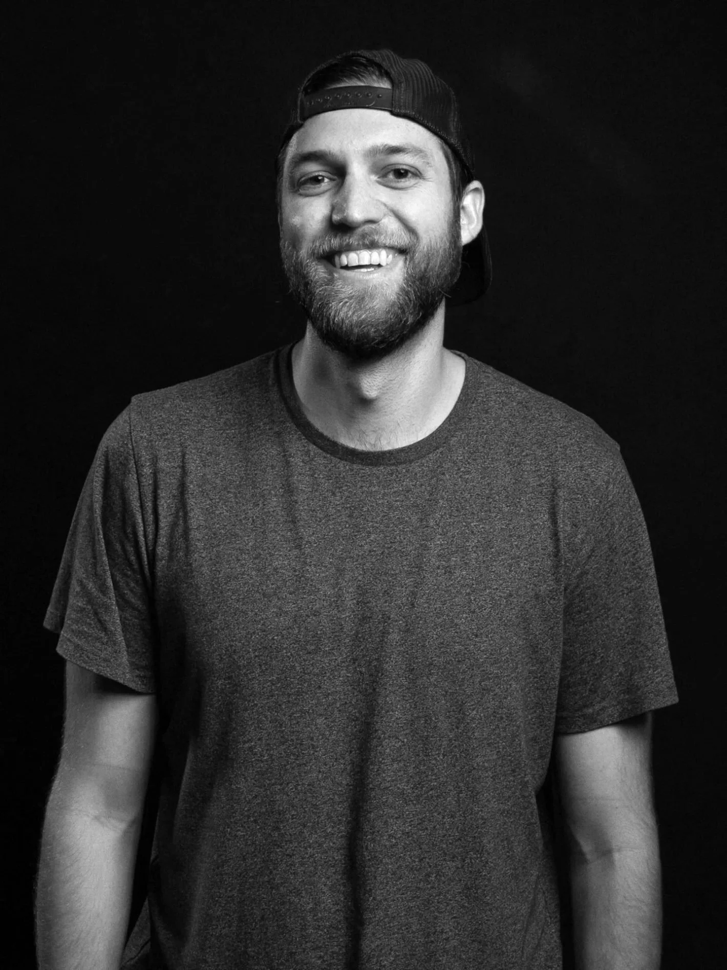 A young man with a beard and mustache wearing a backward baseball cap and a casual t-shirt, smiling against a plain dark background.