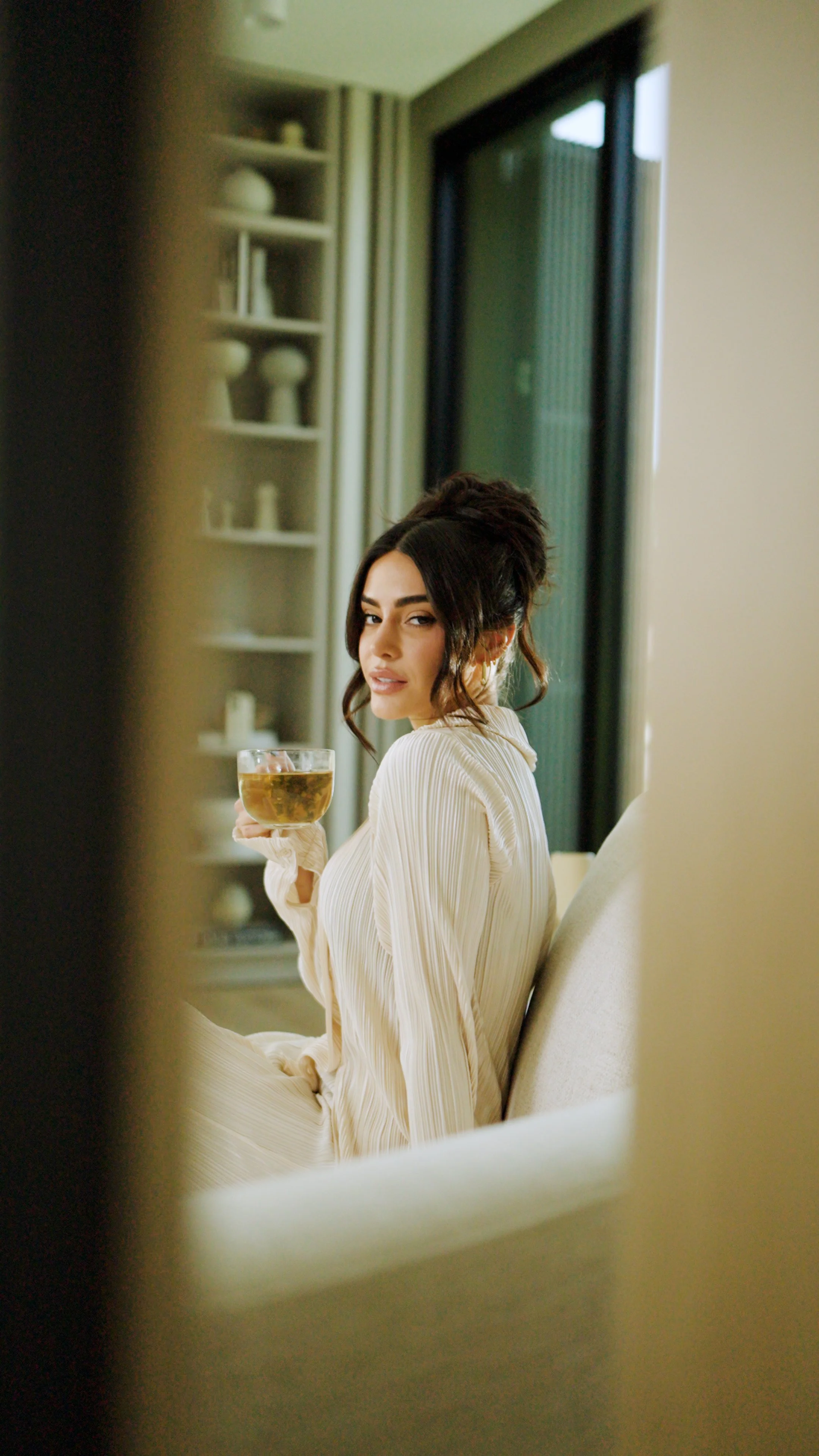 A woman with dark hair in a bun wearing a cream-colored silky dress, sitting on a sofa, holding a glass of tea, looking at the camera.
