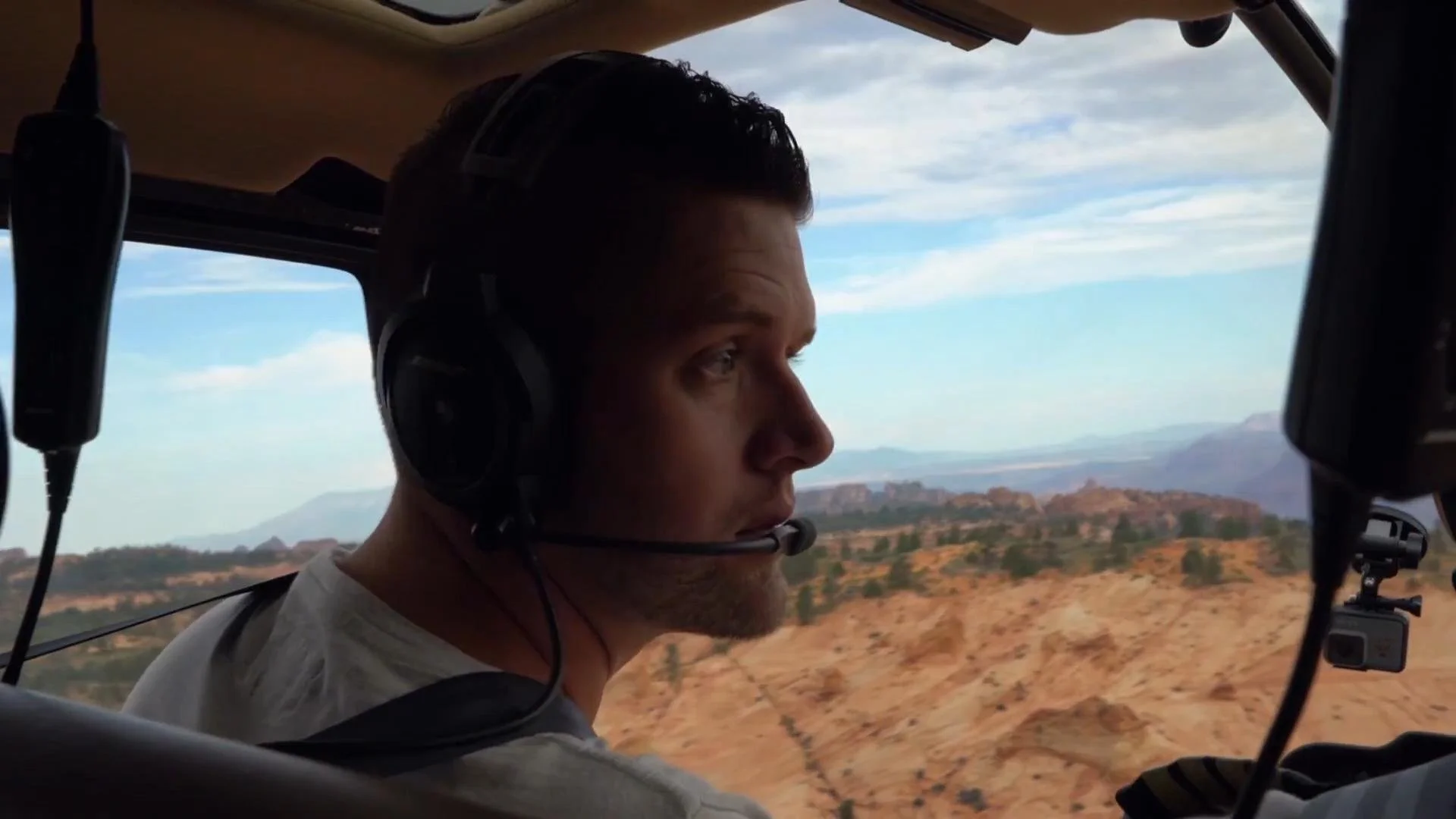 A man wearing a headset sitting inside a helicopter, flying over a desert landscape with mountains in the background.