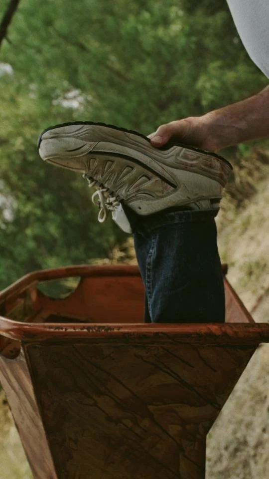 Person holding a sneaker while standing in a wooden box outdoors with green trees in the background.