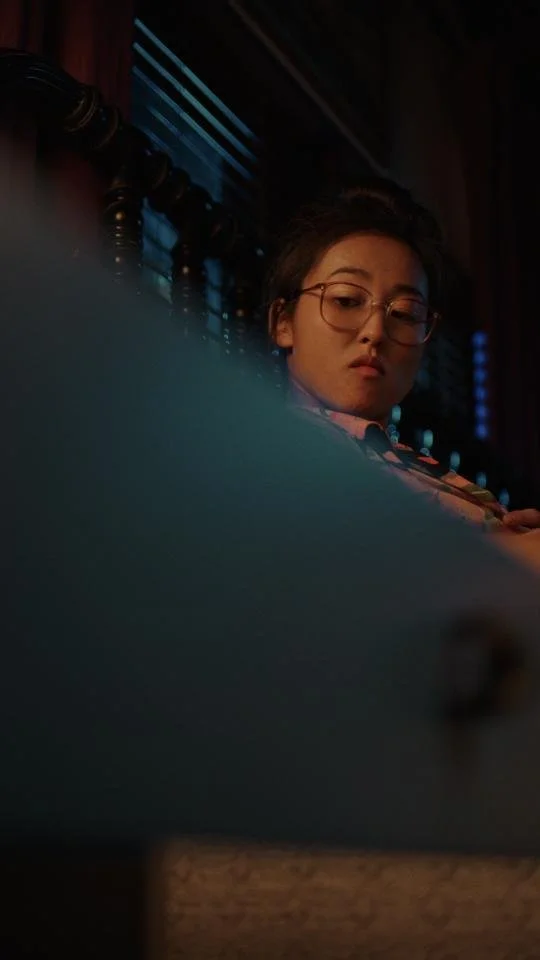 A young woman with glasses looks down, sitting indoors at night with blue lighting and window blinds in the background.