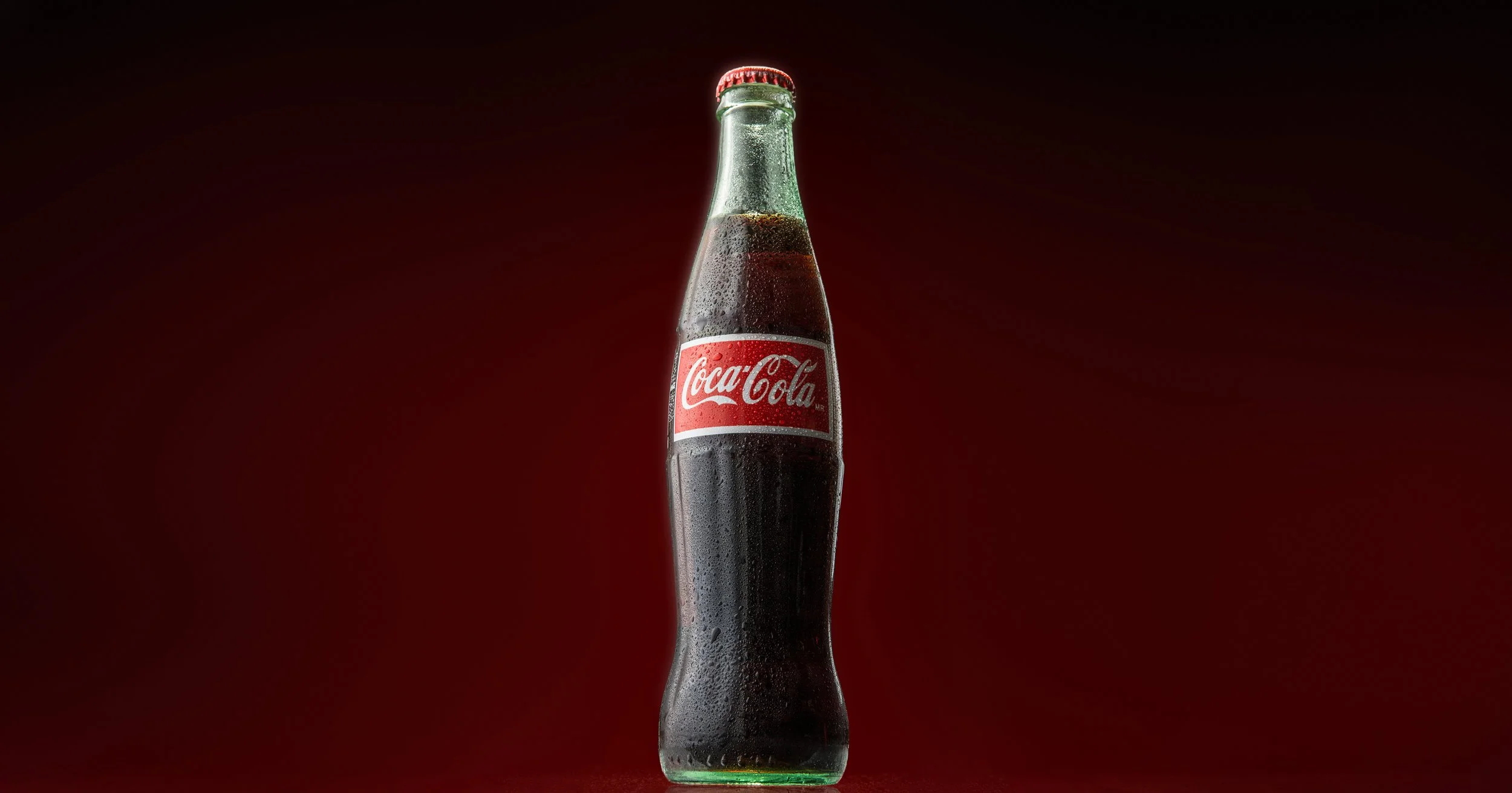 A glass Coca-Cola bottle with condensation and a red label, standing upright against a dark background.