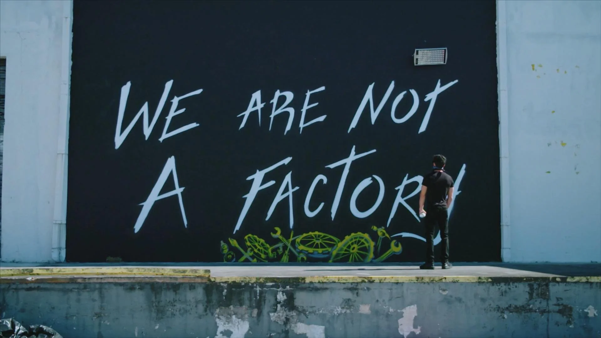 A large mural on a building wall with the text 'We are not a factory' in bold white letters on a black background, featuring a yellow industrial gear graphic at the bottom, and a person wearing black standing in front of it.
