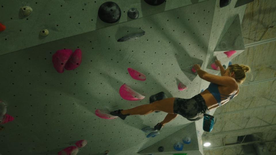 A woman with blonde hair tied in a bun, wearing a blue sports bra and black shorts, is climbing an indoor rock wall with pink and black holds.
