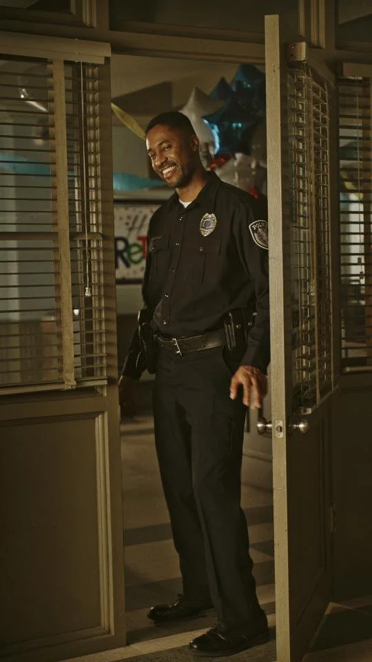 A police officer in uniform standing in an open doorway smiling at the camera.