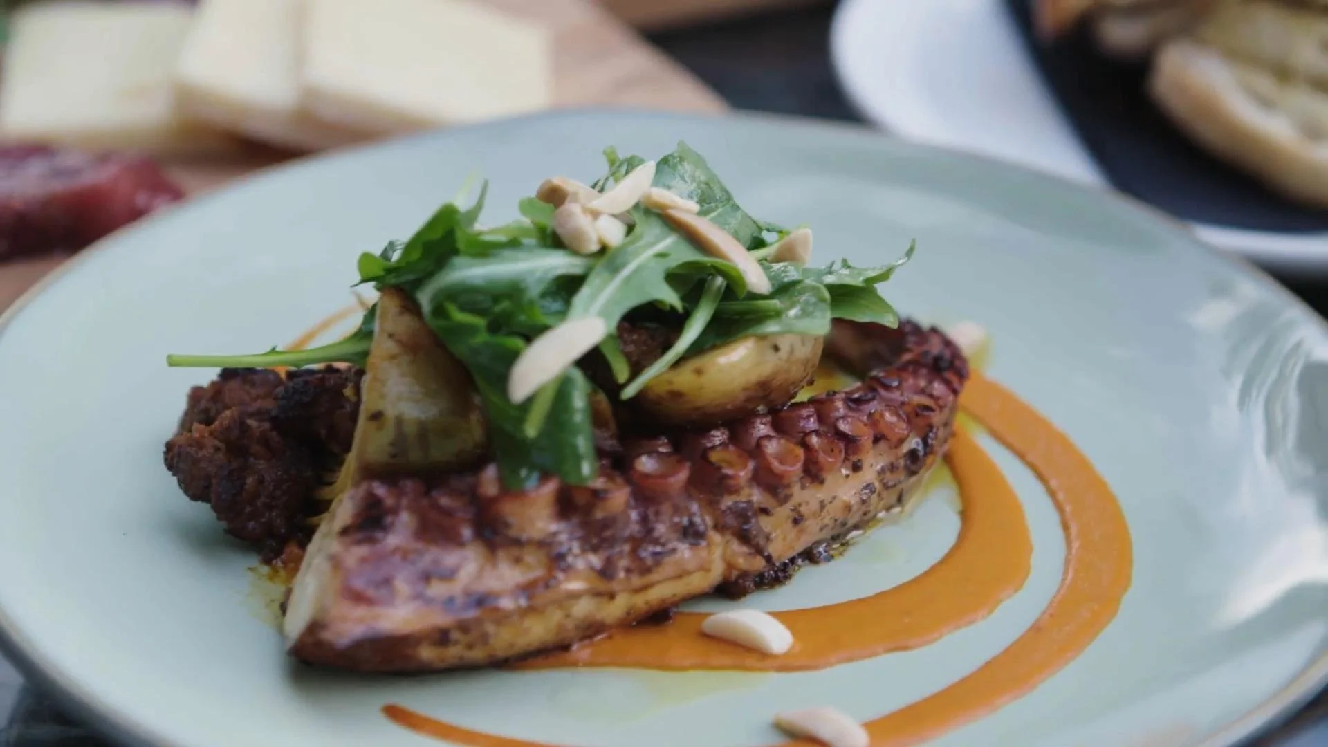 Grilled octopus tentacle topped with mixed greens and slivered almonds, served on a plate with orange sauce decoration.