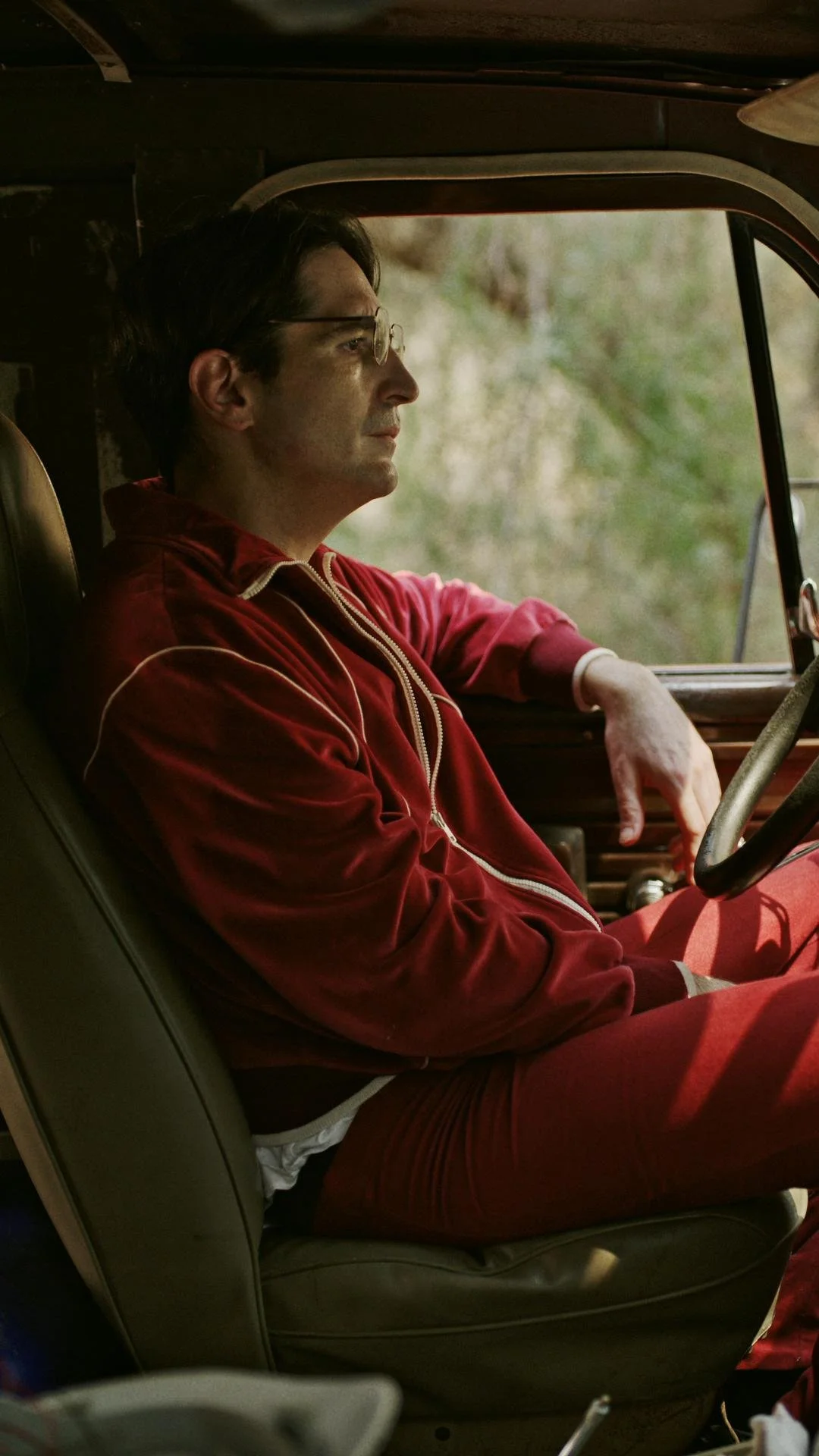 A man sitting in a vehicle with a serious expression, wearing glasses and a red tracksuit, looking out the window.