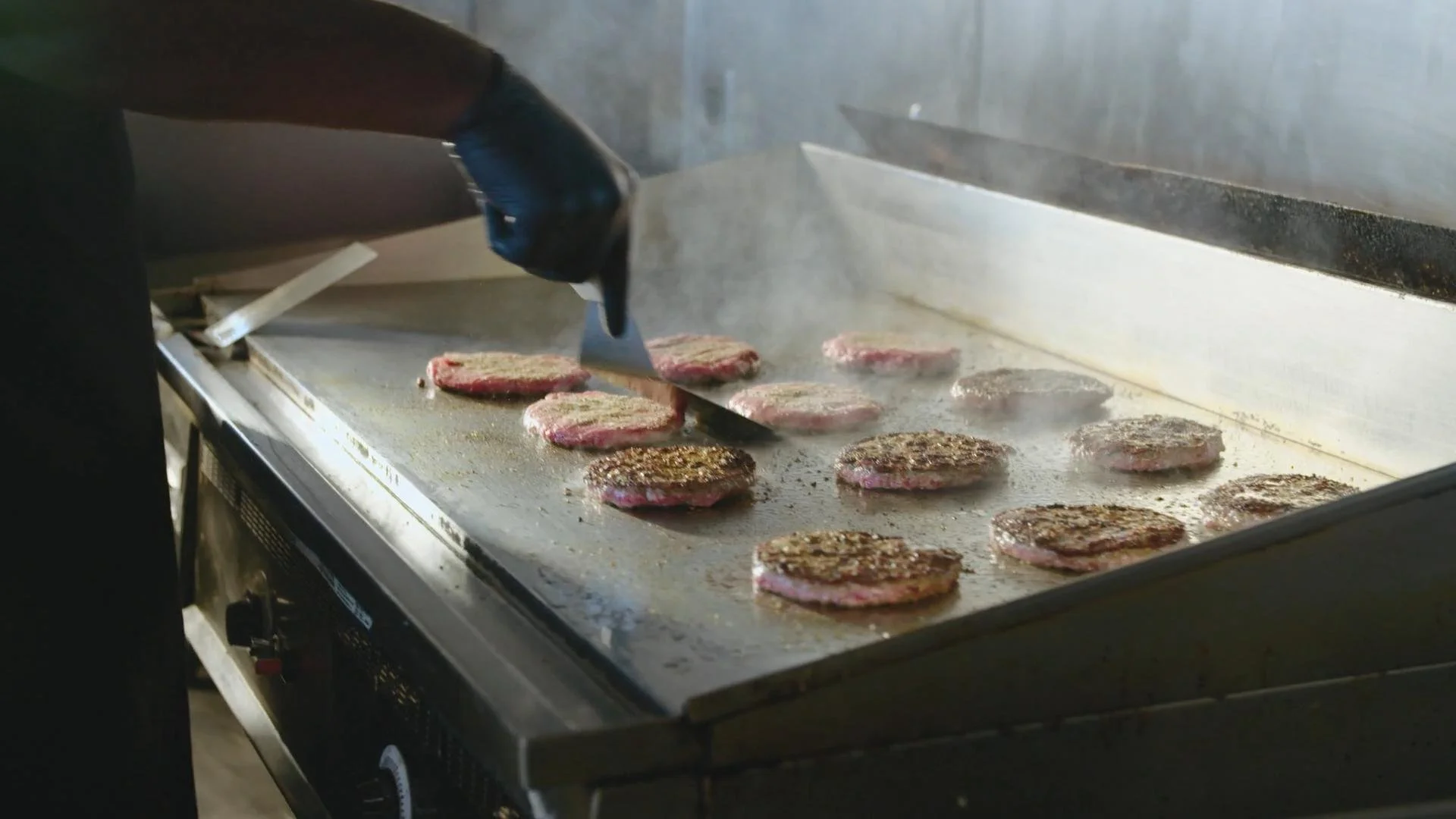 A person wearing black gloves cooking hamburger patties on an electric flat-top grill.