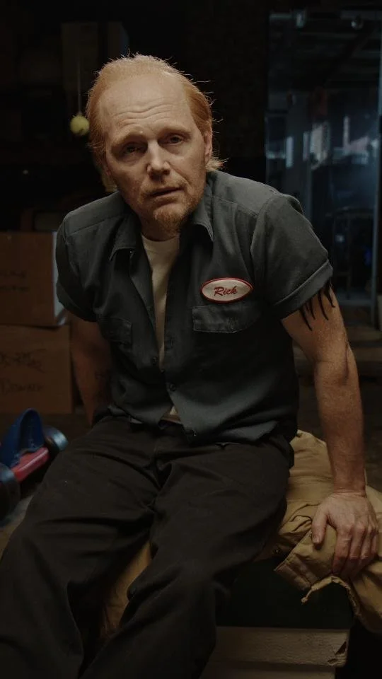 A man with pale skin, reddish hair, and a serious expression, wearing a dark mechanic uniform with a name tag that says 'Rick', sitting on a bench in a dimly lit workshop or garage, with tools and boxes around.