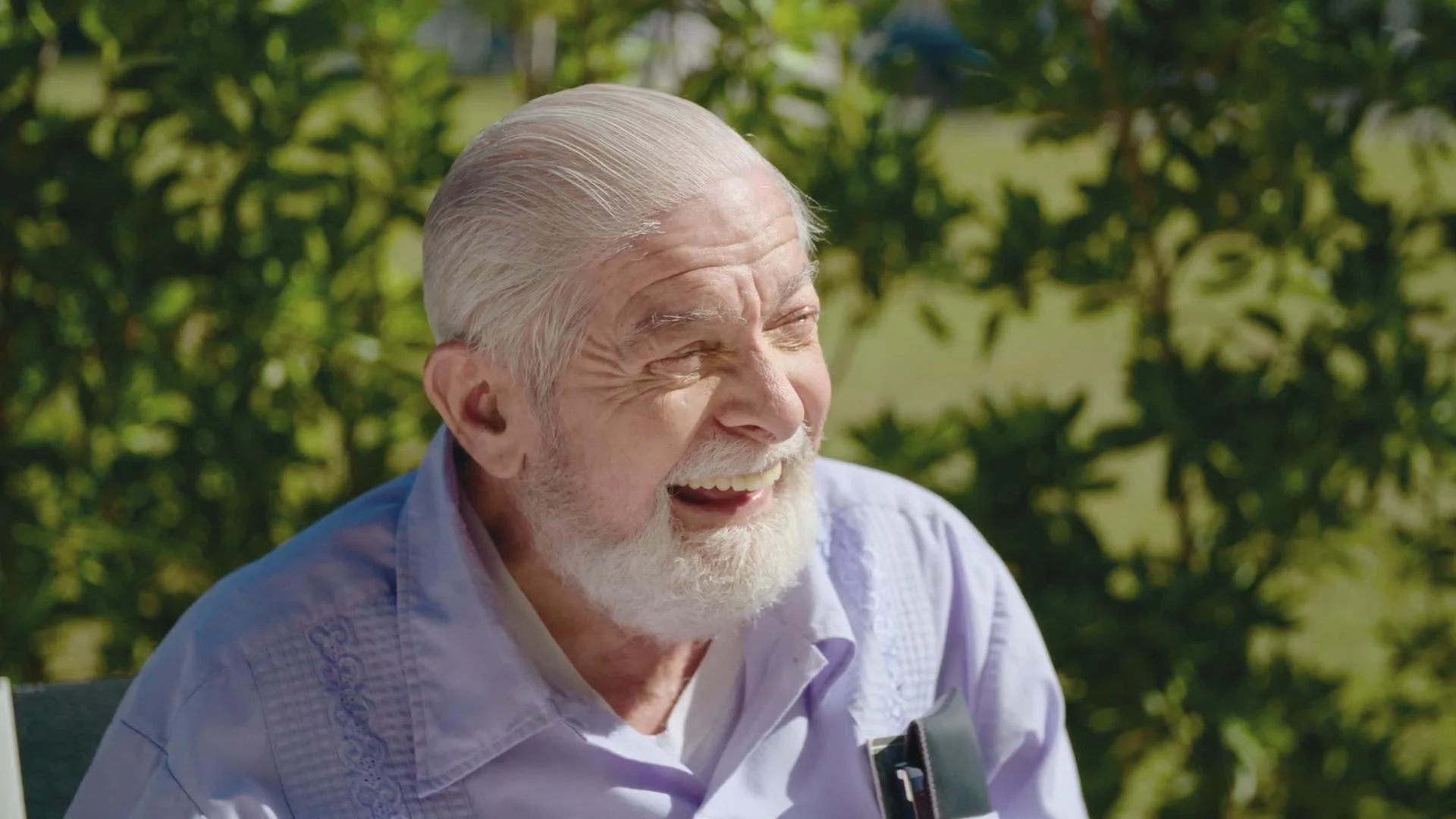 An elderly man with a white beard and silver hair smiling outdoors, wearing a light purple shirt with embroidery, with green trees in the background.