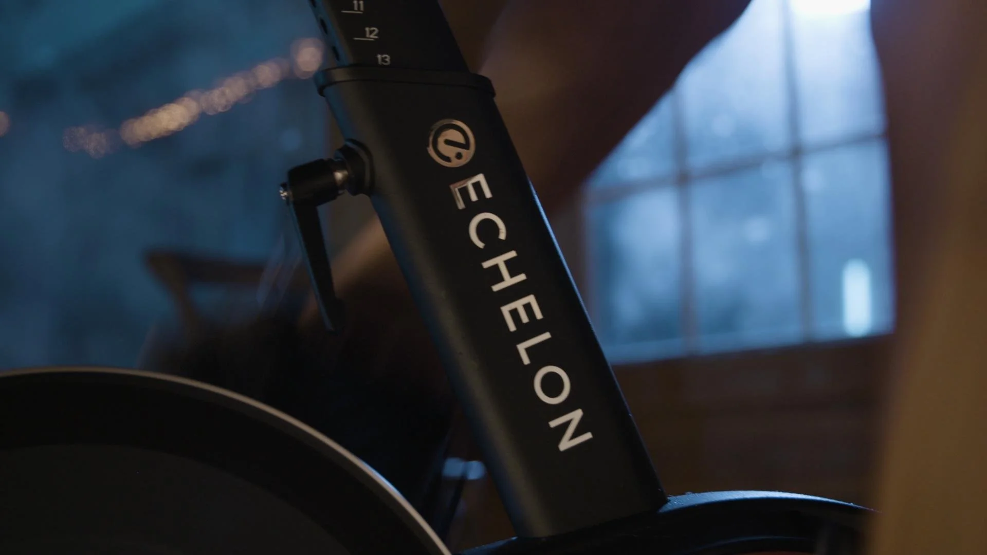 Close-up of a black Echelon stationary exercise bike with the brand name visible, set in a room with a window allowing natural light to come through.