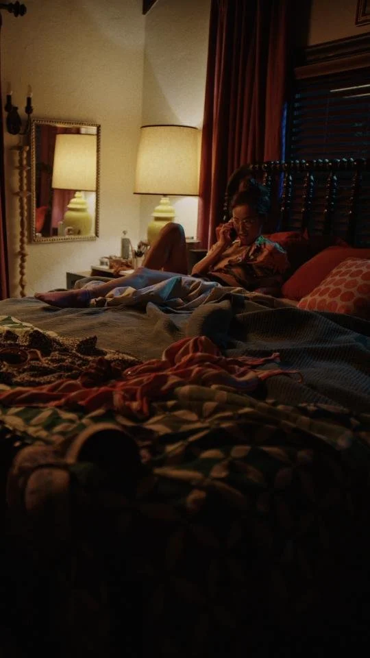A woman sitting on a bed in a dimly lit bedroom, talking on the phone. The bed has blankets and pillows, and there are lamps and a mirror behind her.
