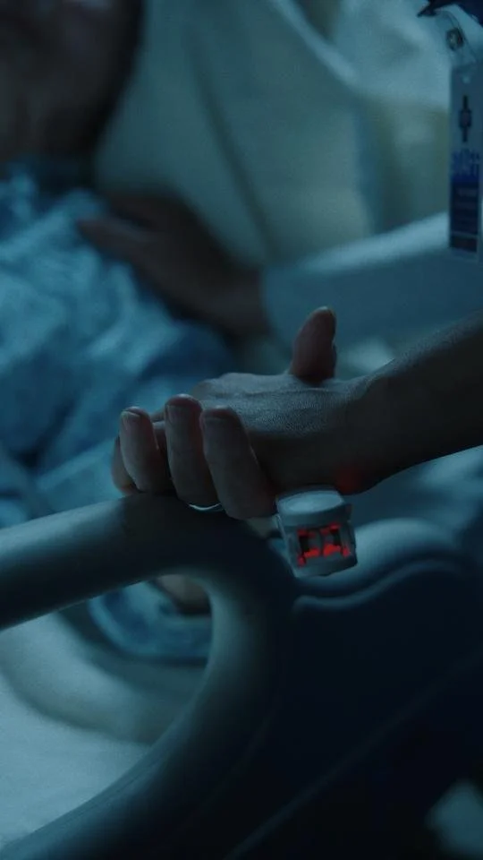 Close-up of a person's hand with an IV attached, resting on a hospital bed rail in a dimly lit hospital room.