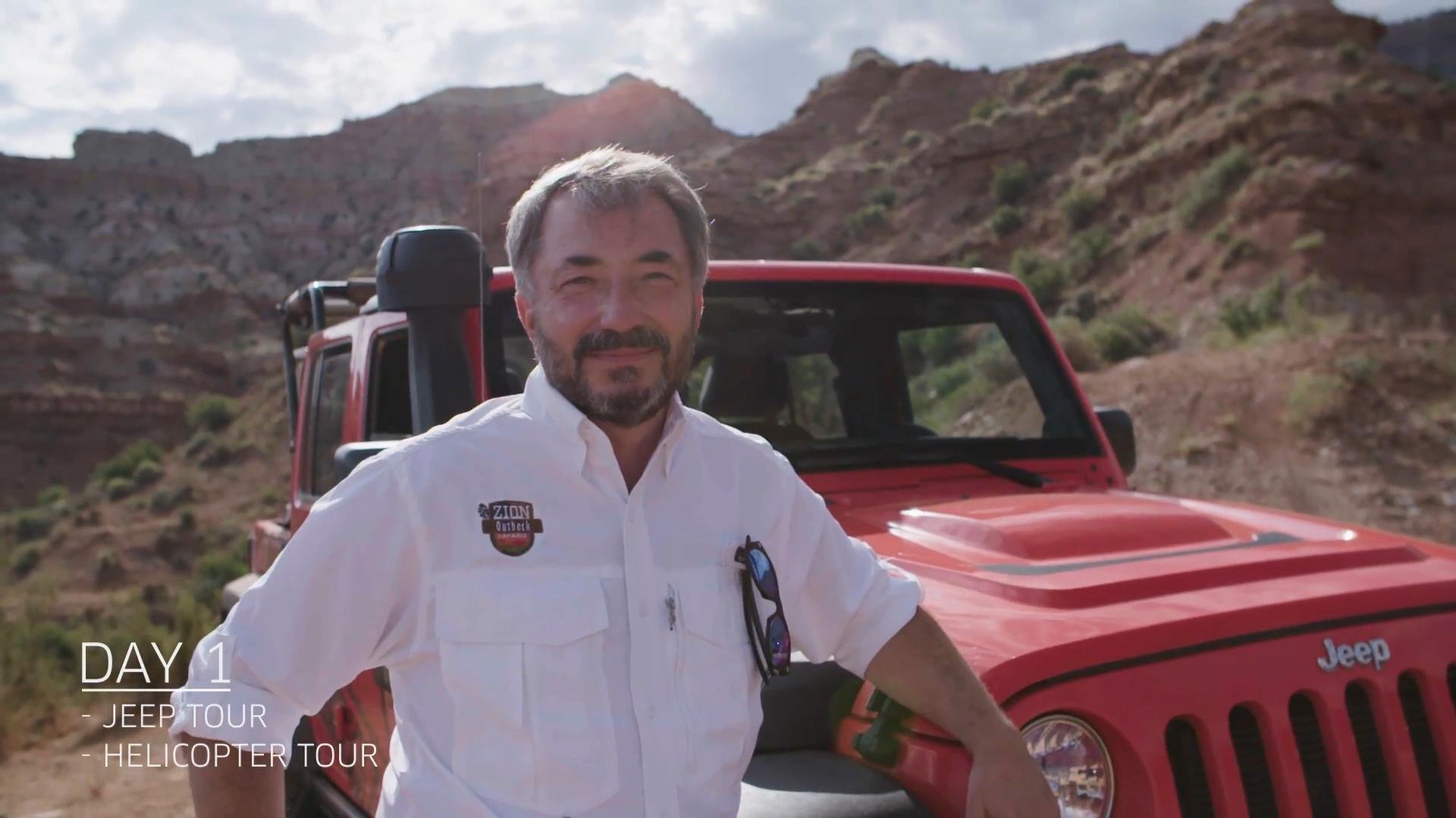 A man smiling outdoors in front of a red Jeep vehicle during a daytime helicopter tour in a desert landscape with rocky hills in the background.