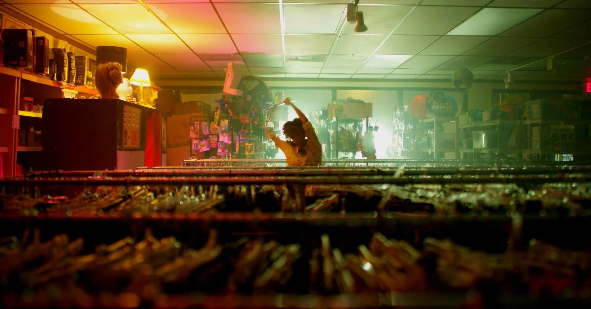 A person with curly hair in a checkered shirt dancing with a stick in a colorful room filled with shelves, toys, and bright lighting.